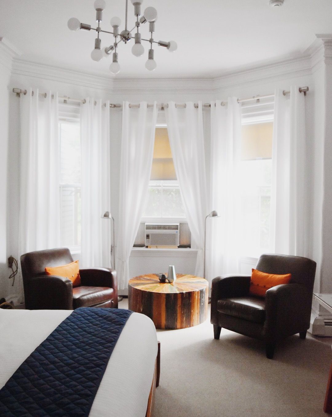 Two dark leather armchairs with orange pillows face a round wooden stump coffee table in a bright, white windowed room.