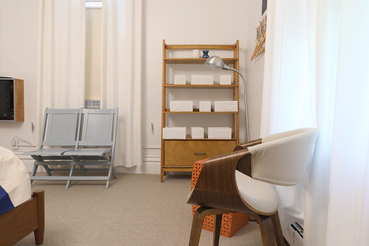 A modern bedroom with white walls, a wooden shelving unit, a grey double chair, and an armchair by a white curtain.