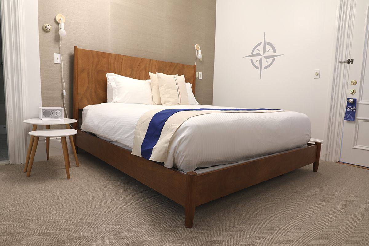 A neatly made bed with a brown wood frame, white linens, and a blue-and-cream runner, set against a grey-textured wall.