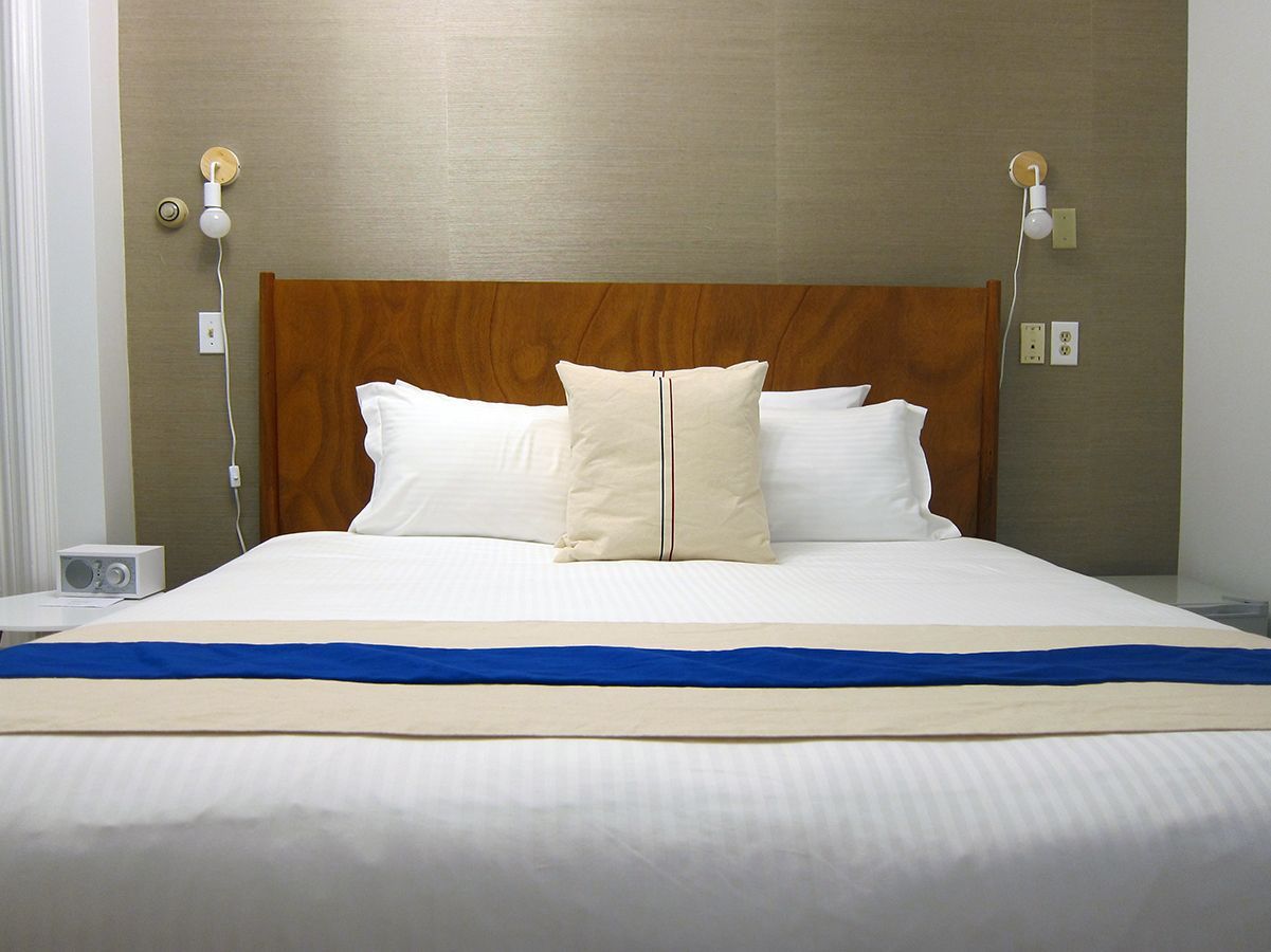 A neatly made bed with white linens, a central tan throw pillow, and a long blue runner against a wooden headboard.
