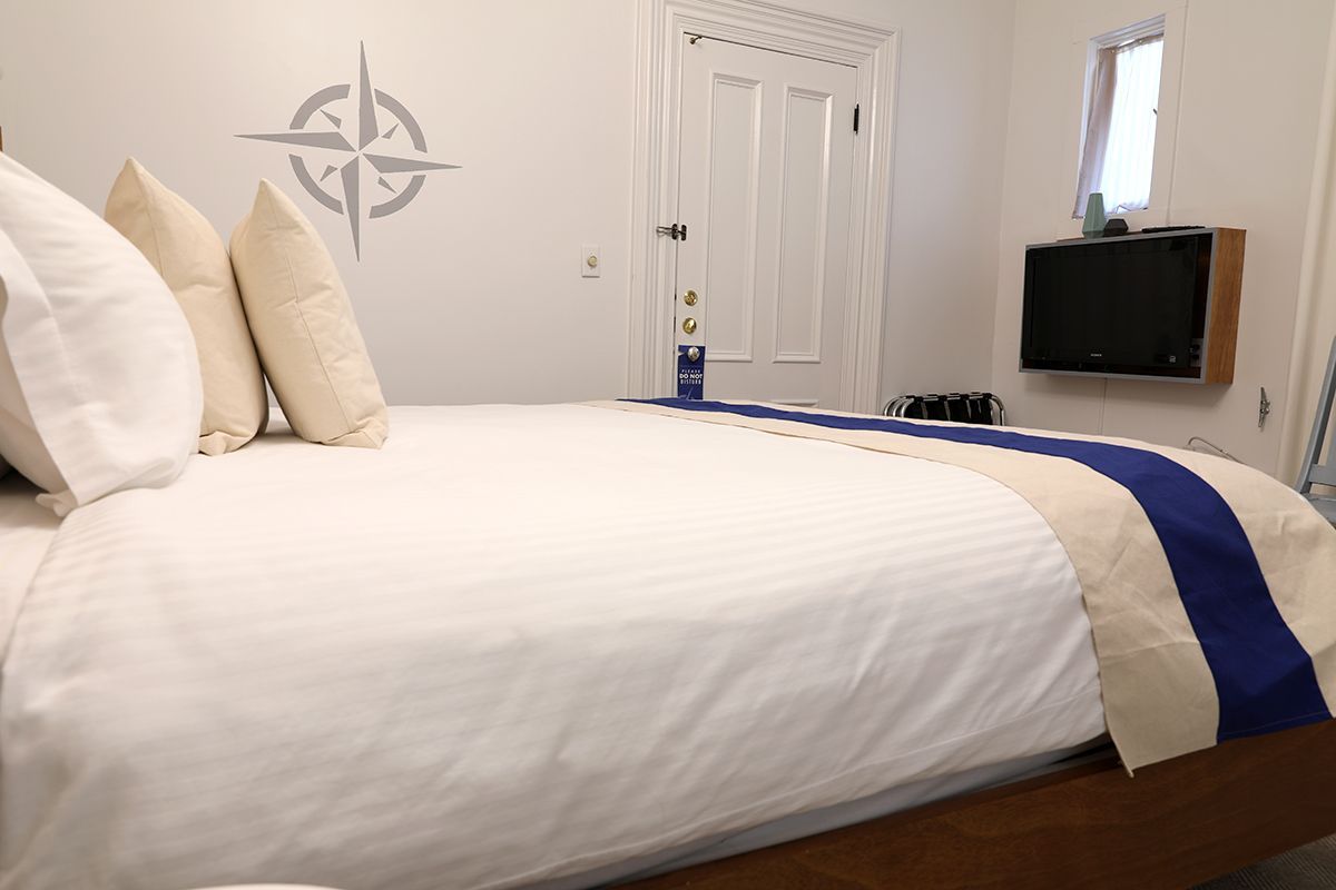 A hotel bed with crisp white linens and a blue runner, featuring a compass wall decal and a wall-mounted TV.