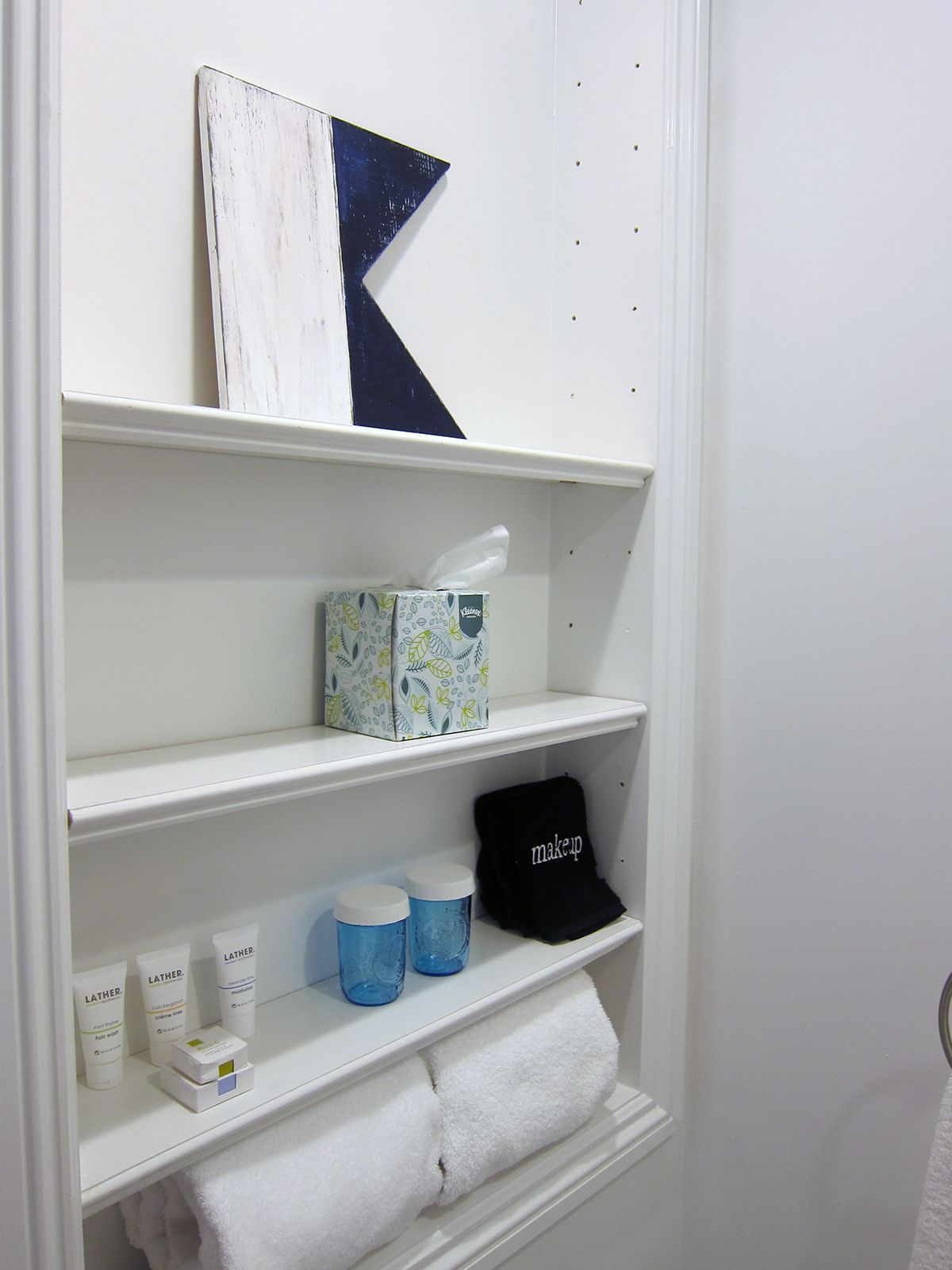 White built-in shelves in a bathroom holding a decorative sign, tissues, toiletries, and folded white towels.