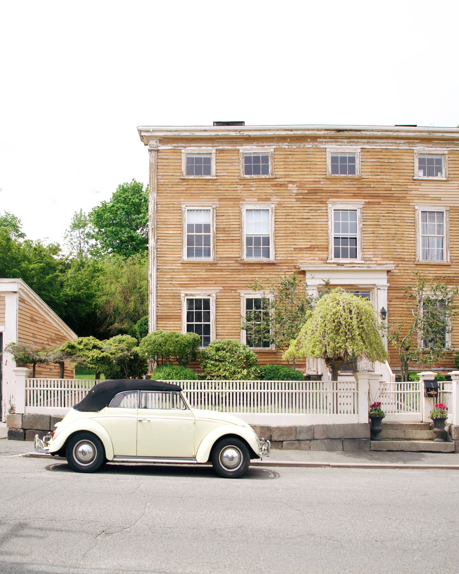 A vintage cream-colored convertible parked on a street in front of a rustic, three-story wooden house with white trim.