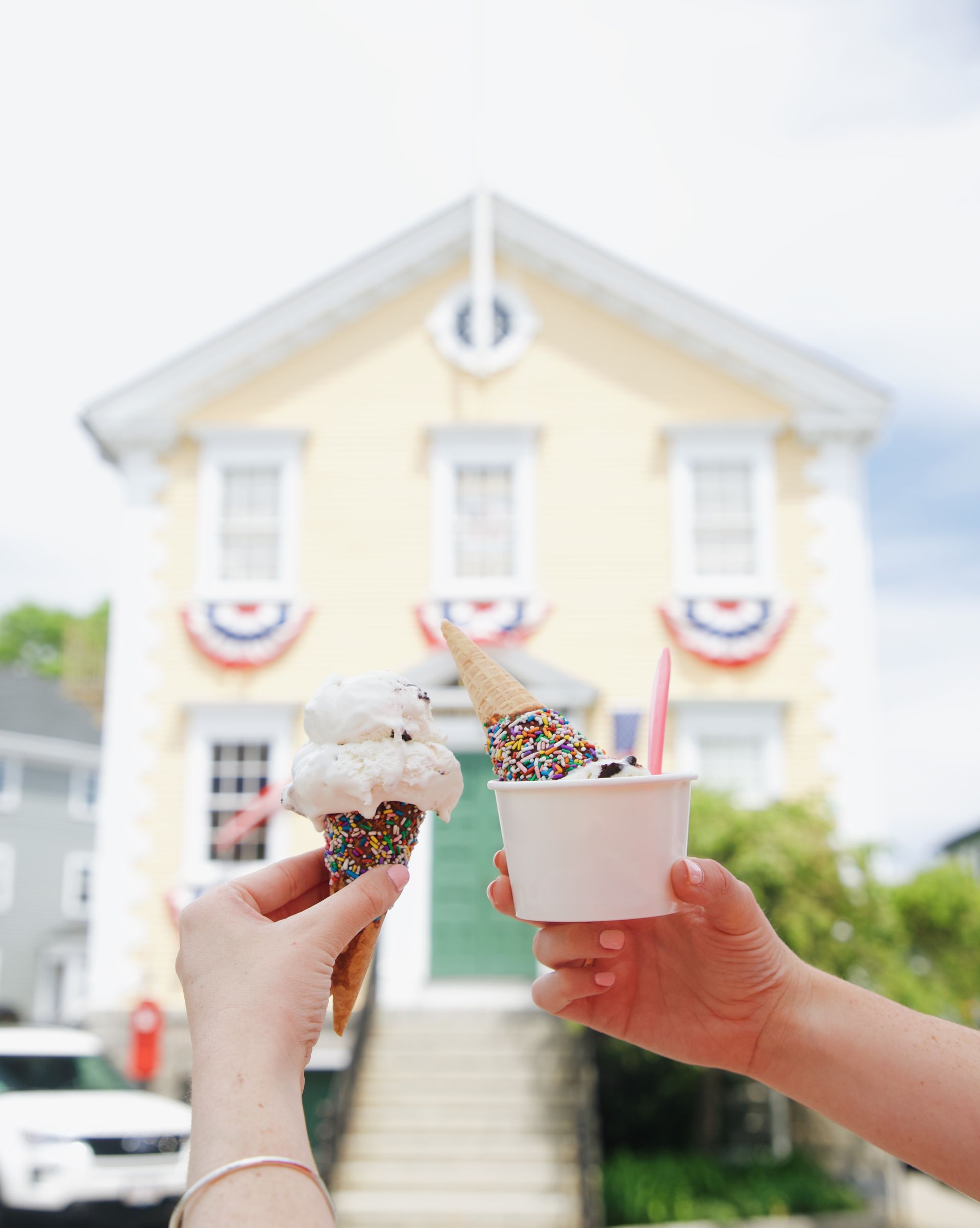 Two hands hold ice cream cones and a cup in front of a yellow building decorated with patriotic bunting.
