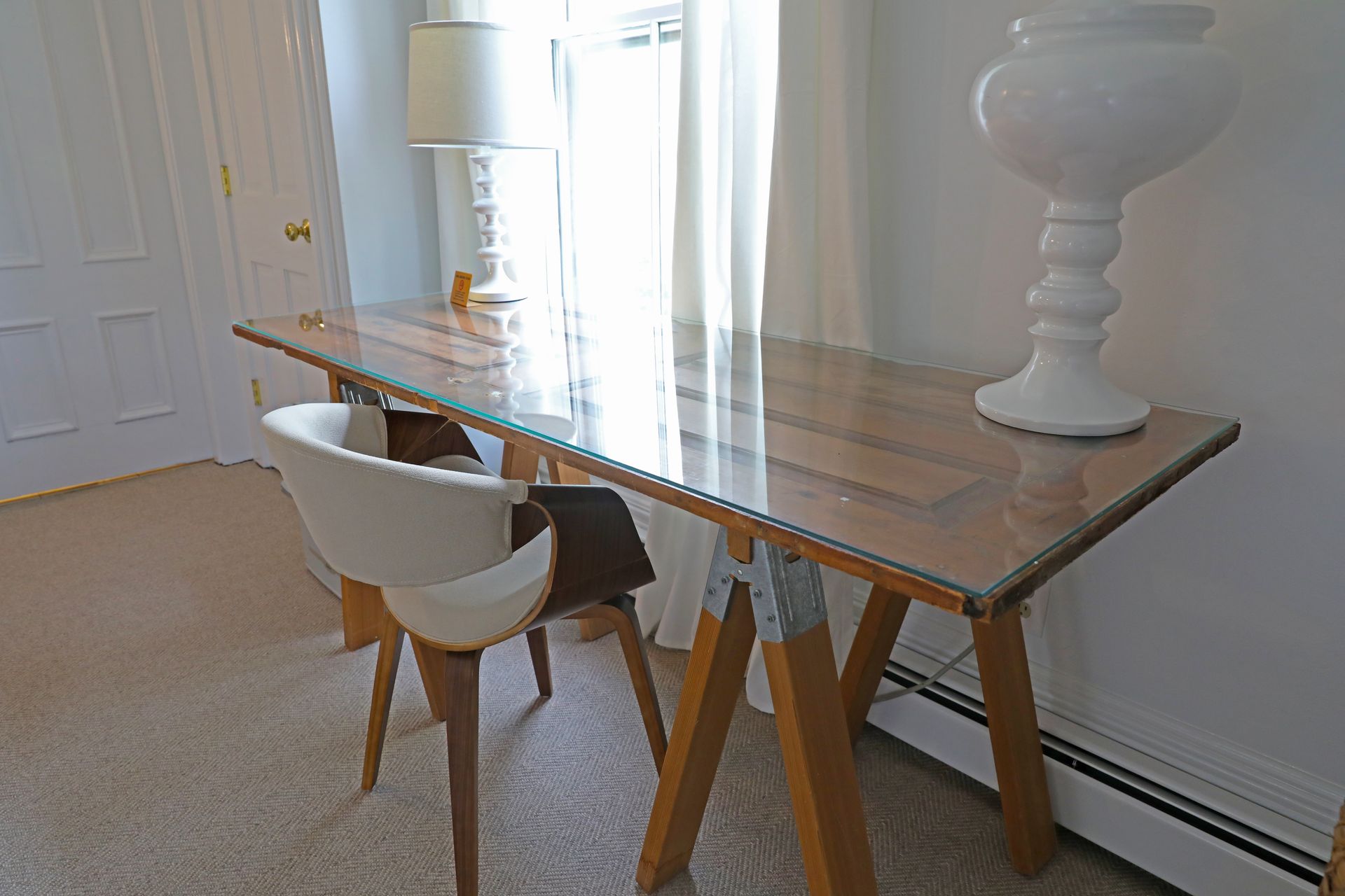 A wooden desk with a glass top and industrial-style legs, paired with a white chair in a bright room.