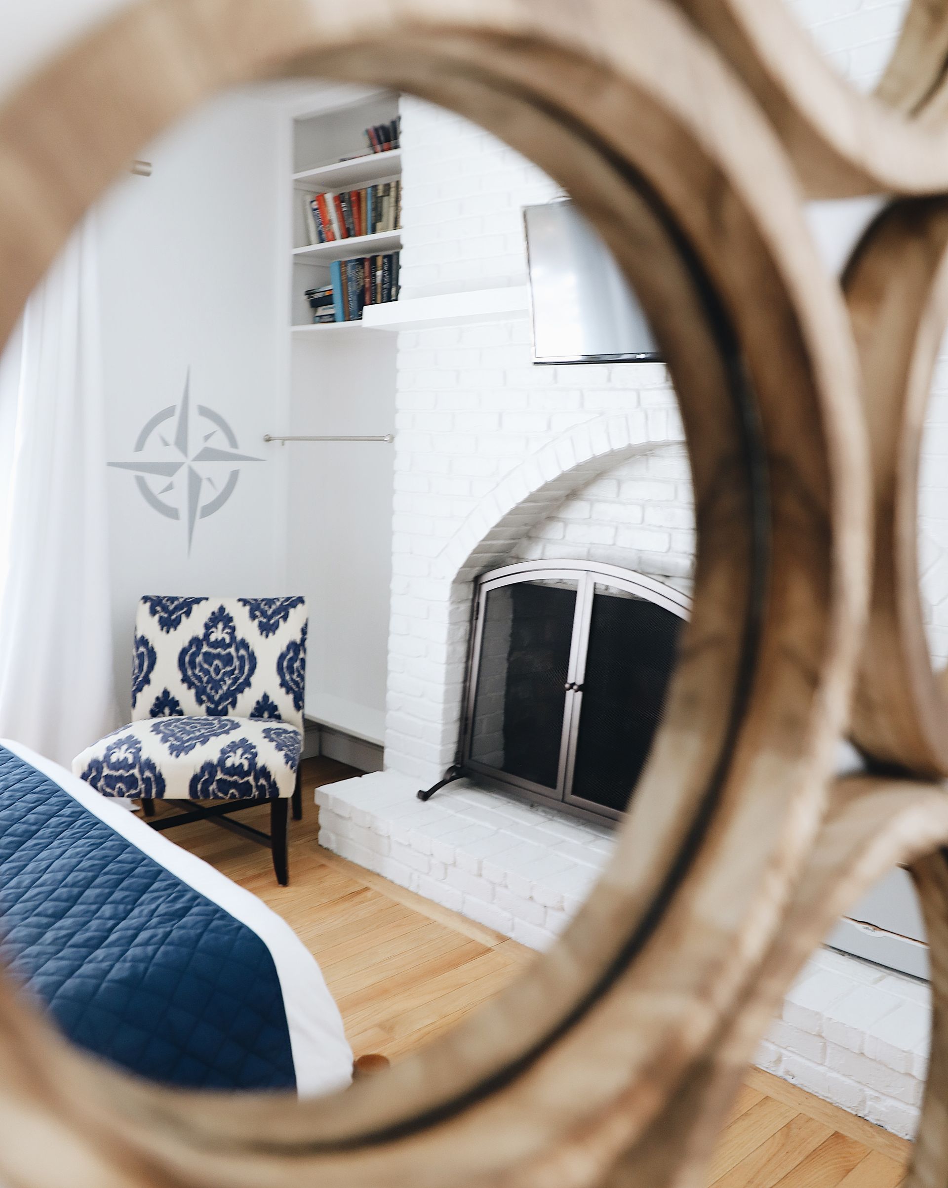 A view through a wooden mirror frame showing a white fireplace, an armchair with blue patterned fabric, and a bed.