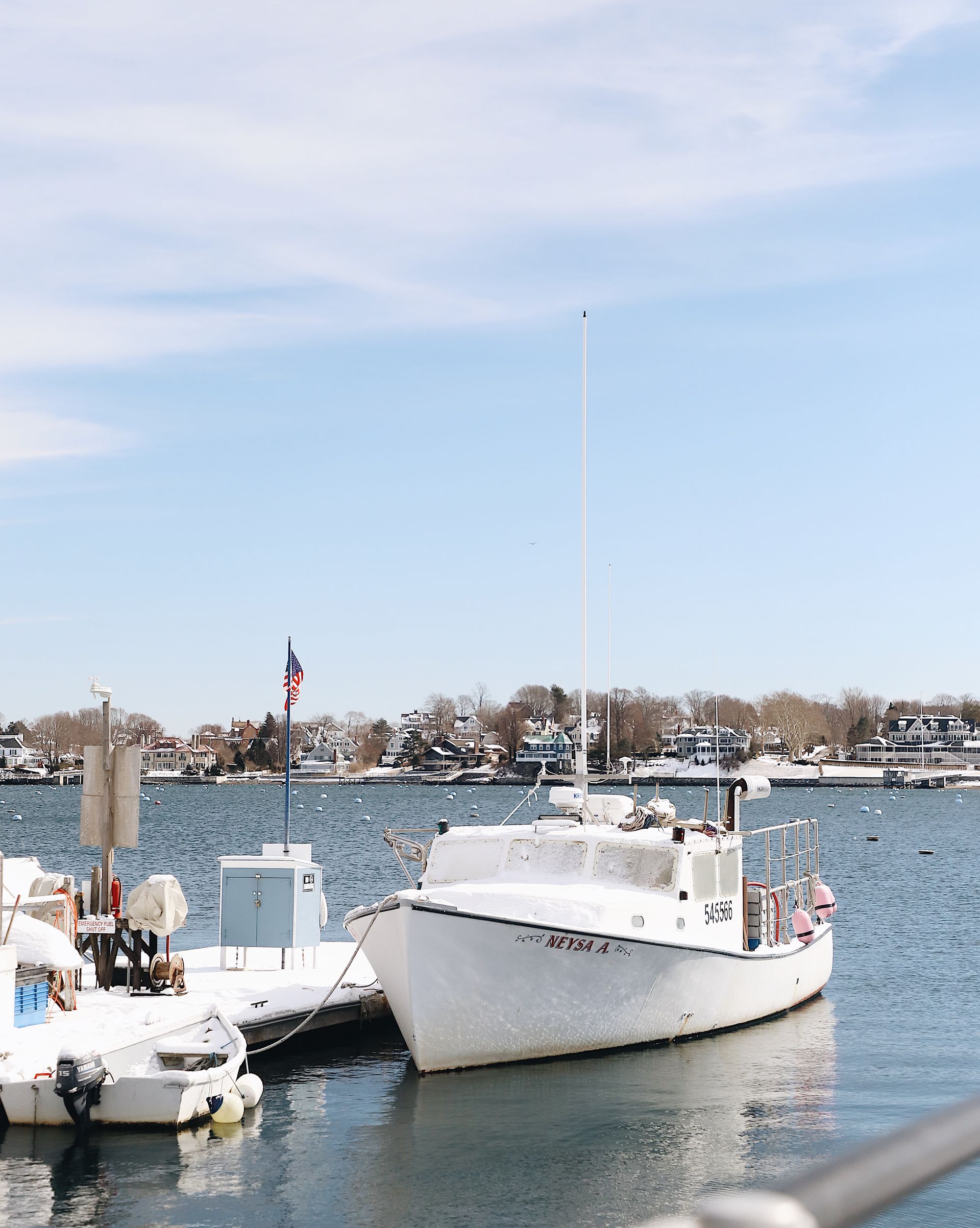 A white fishing boat moored at a dock on a sunny day with trees and houses along the distant shoreline.