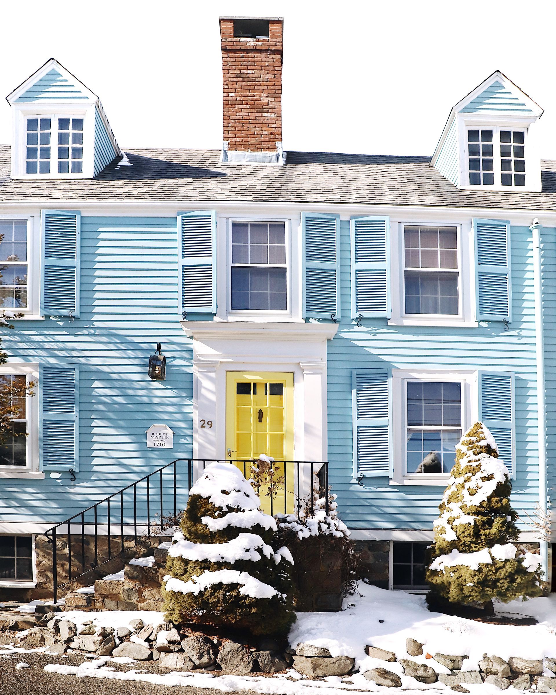 A light blue, two-story wooden house with a bright yellow front door, brick chimney, and snow-covered ground.