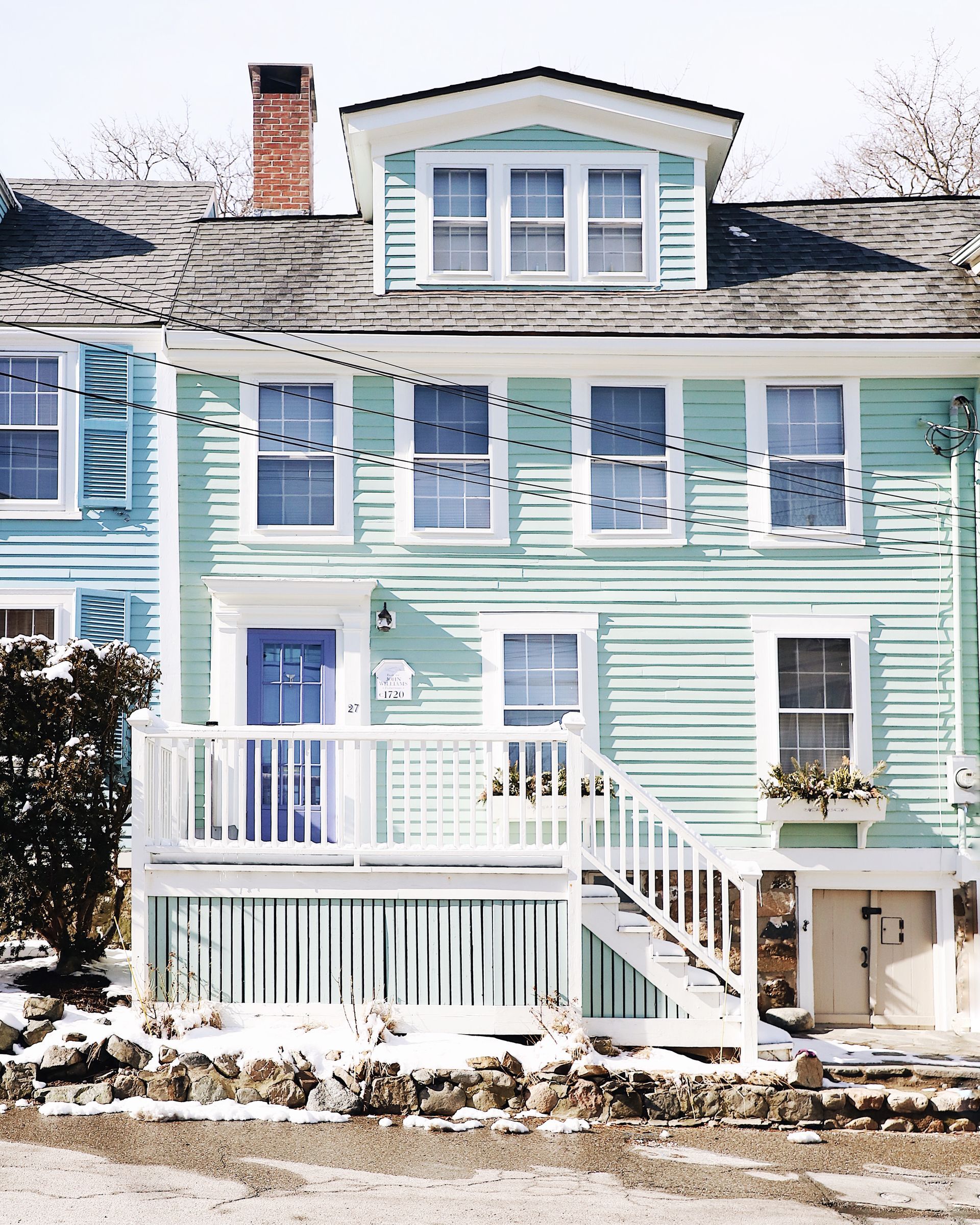A pale green house with white trim, a wooden porch, and a dormer window, viewed on a sunny day with patches of snow.