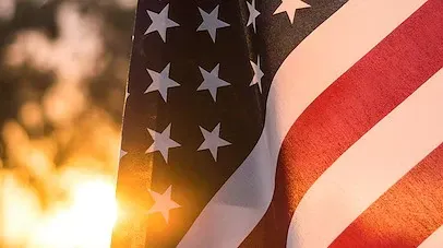 A close-up of an American flag waving in the sunlight, with a soft-focus background of trees.