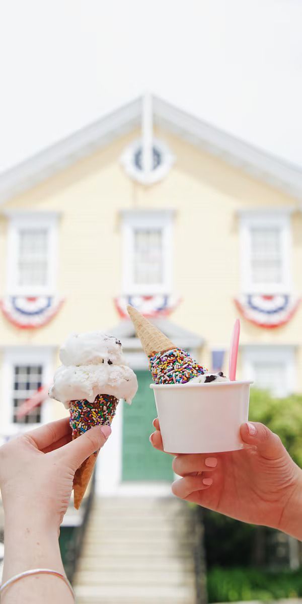 Two hands hold ice cream treats against a blurred background of a yellow colonial-style building with bunting.