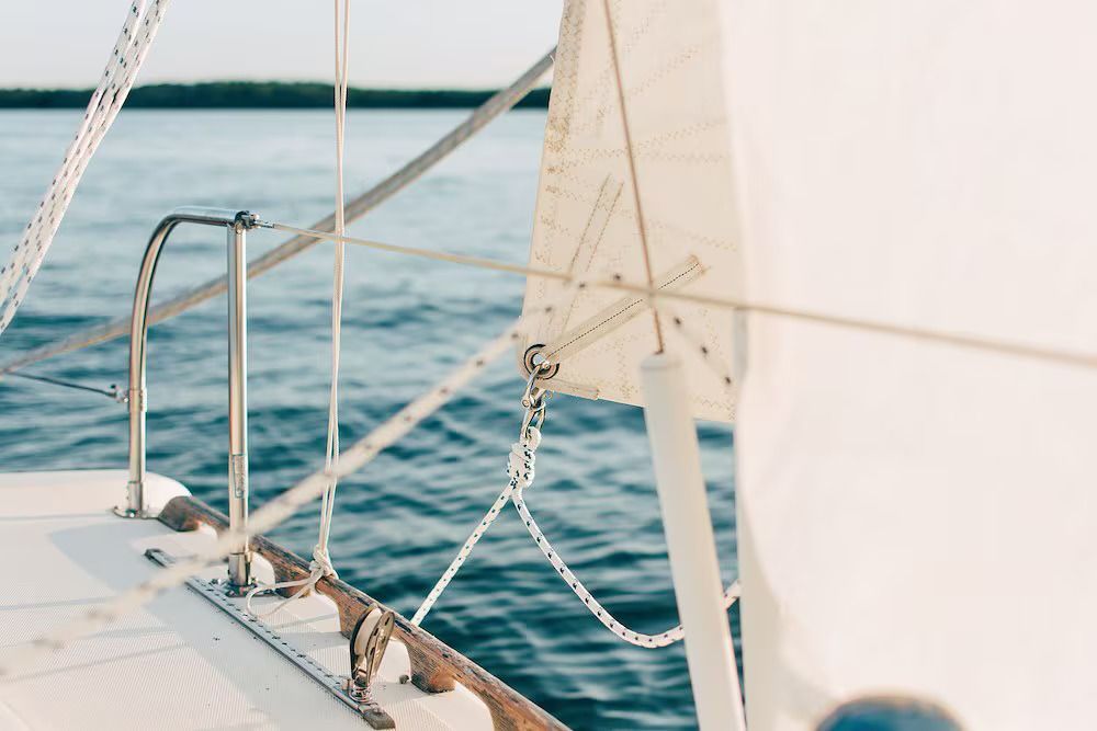 Close-up of a sailboat deck with white sails, metal rigging, and lifelines overlooking blue water and a distant shoreline.
