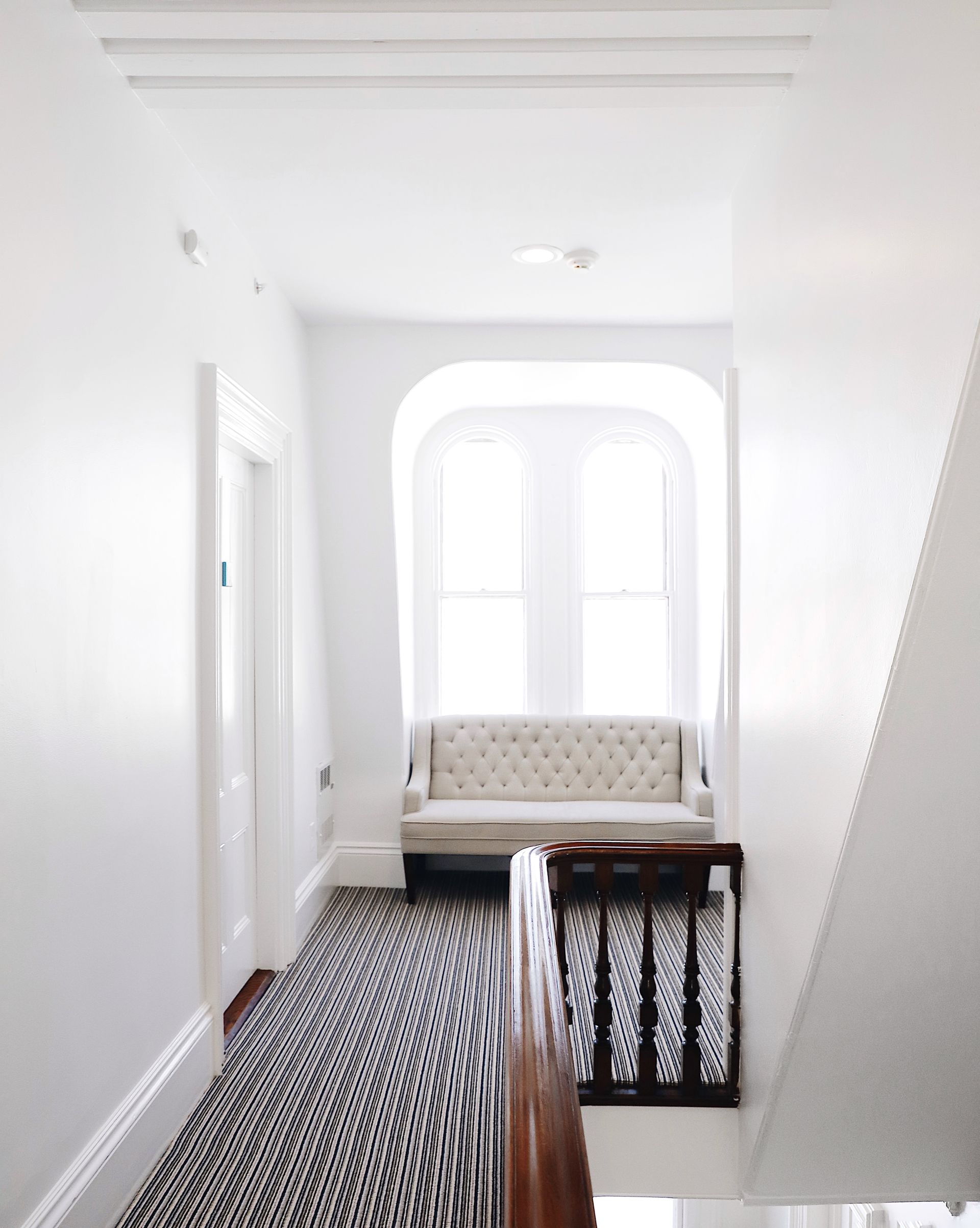 A bright hallway with a black-and-white patterned carpet leading to a small, tufted loveseat set before an arched window.