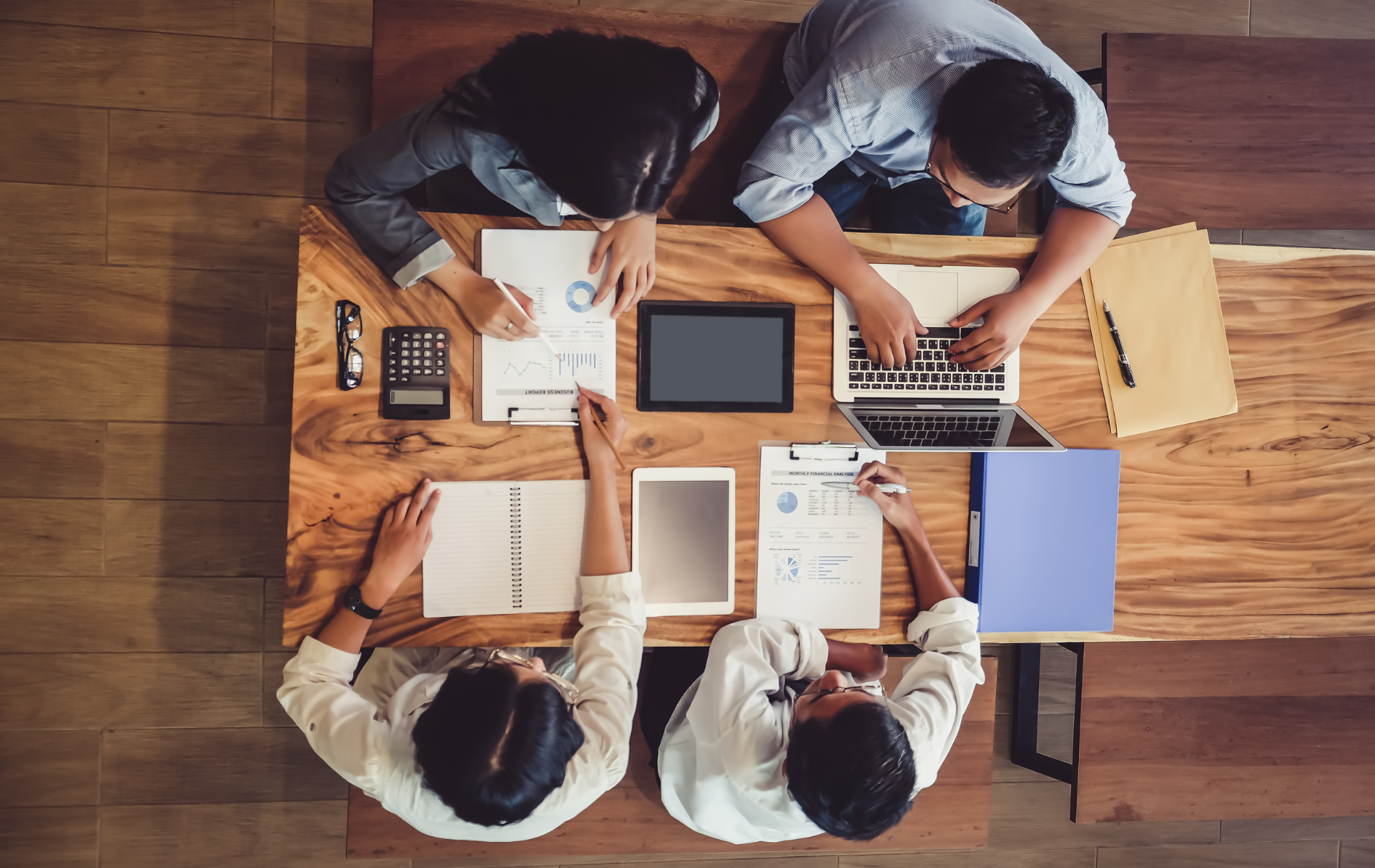 Four people working collaboratively at a wooden table with papers, a laptop, and a tablet.