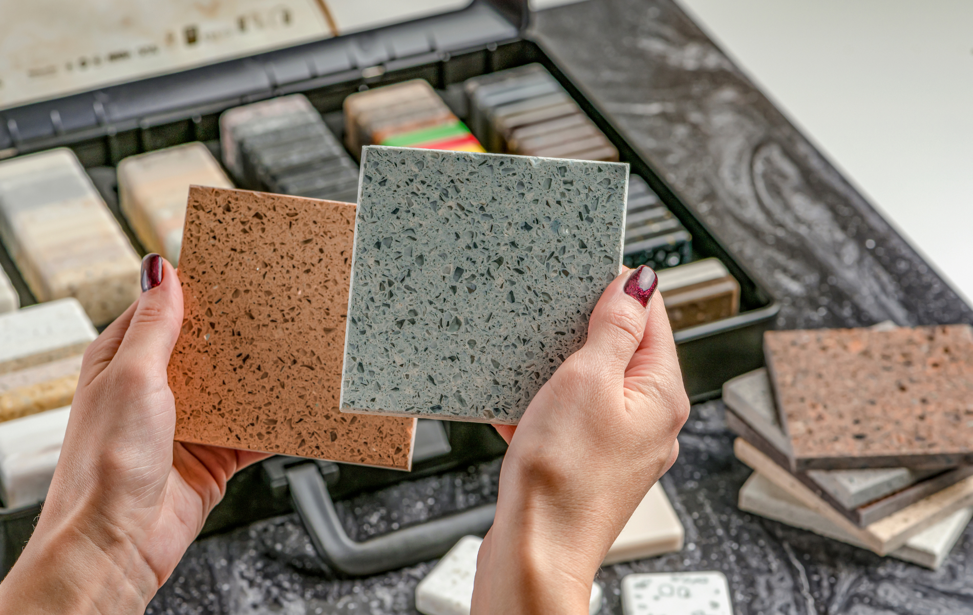 Hands holding brown and blue speckled tiles from a sample case, with other samples in the background.