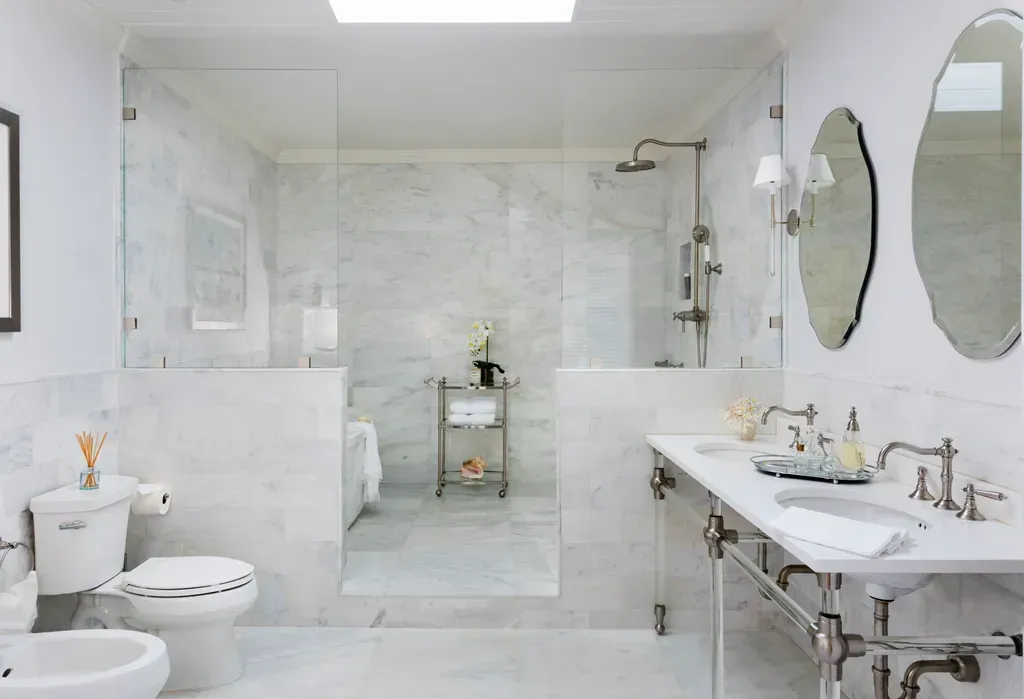 A luxurious, all-white marble bathroom featuring a walk-in glass shower, double vanity, and separate toilet area.