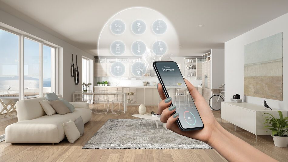 A person's hand holding a phone controlling a smart home. A light-filled living room is in the background.