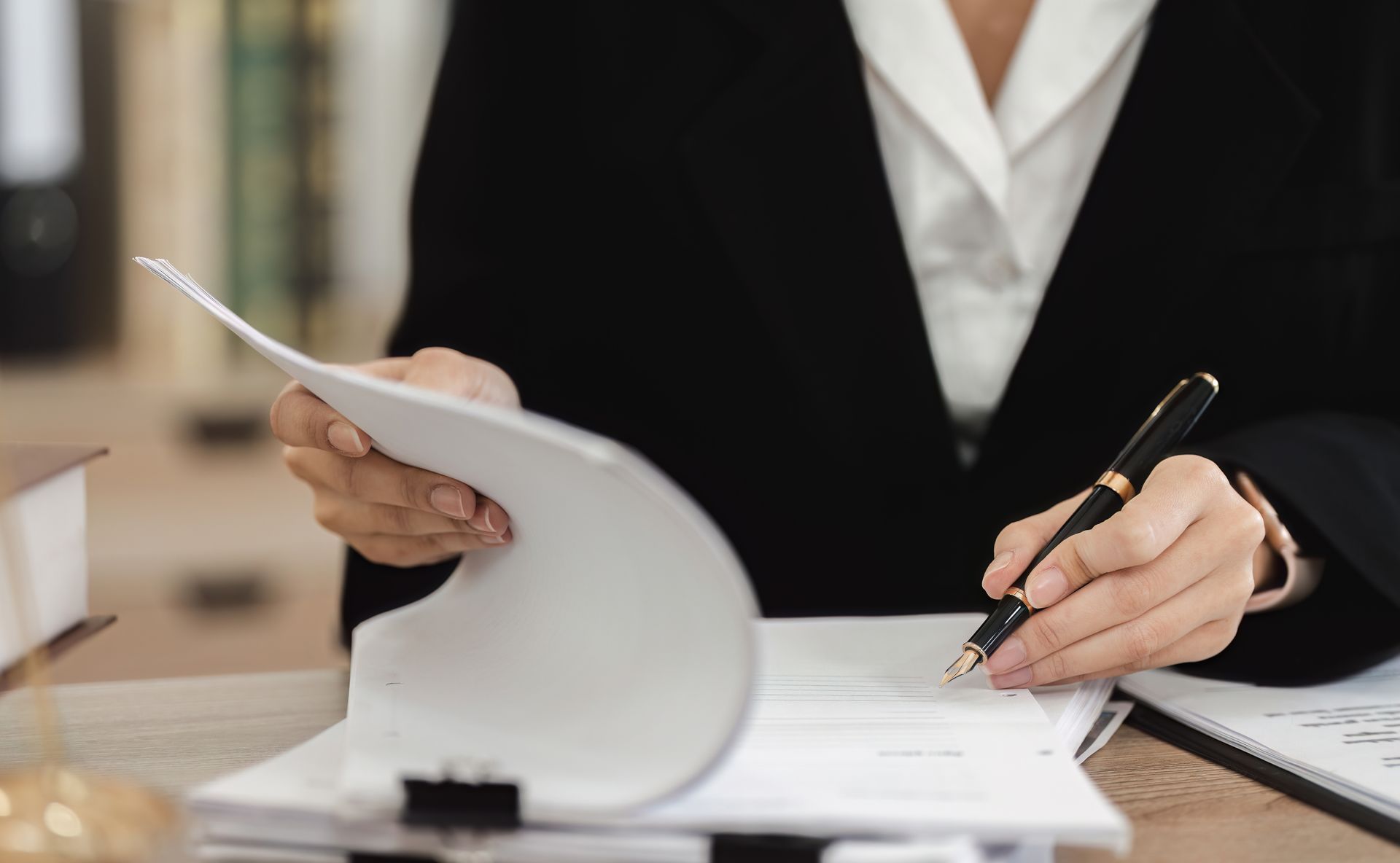 Person in black suit writing on papers at a desk with a pen, in an office setting.
