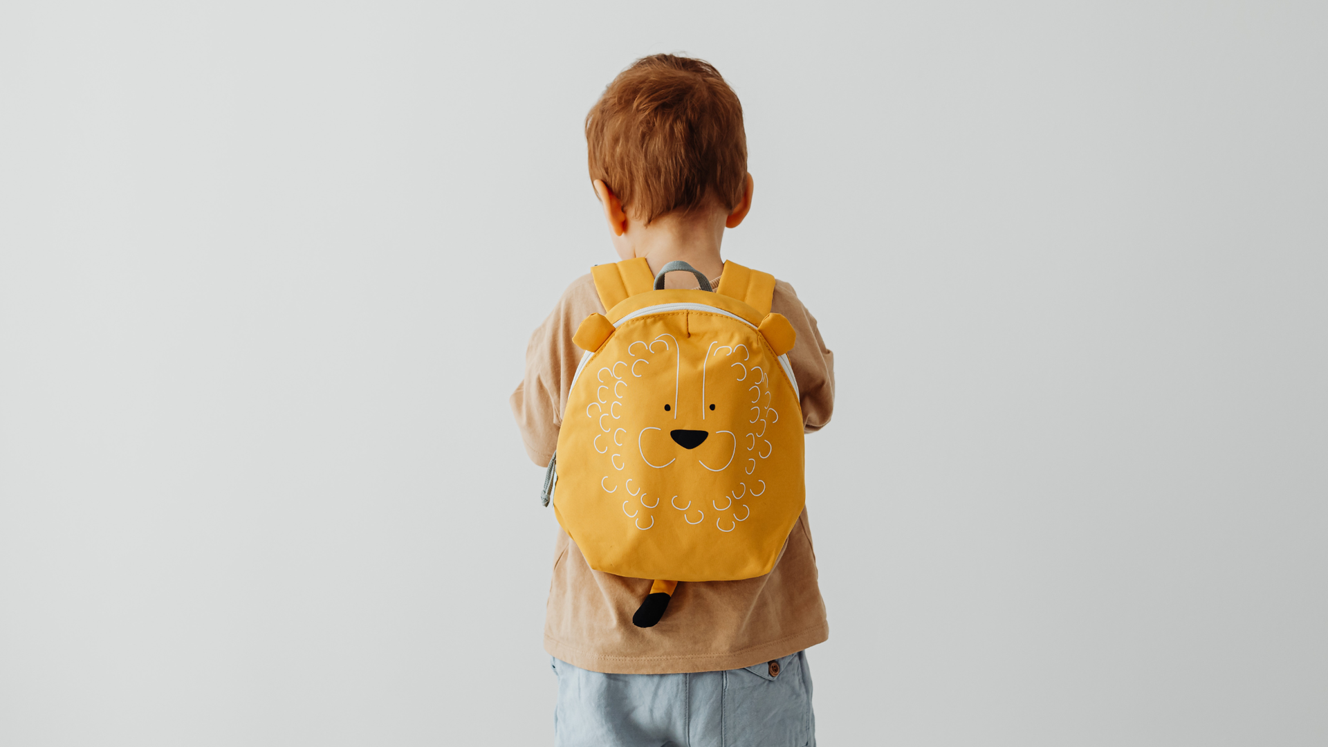 A child seen from behind wearing a tan shirt and a yellow backpack featuring a cartoon animal face.