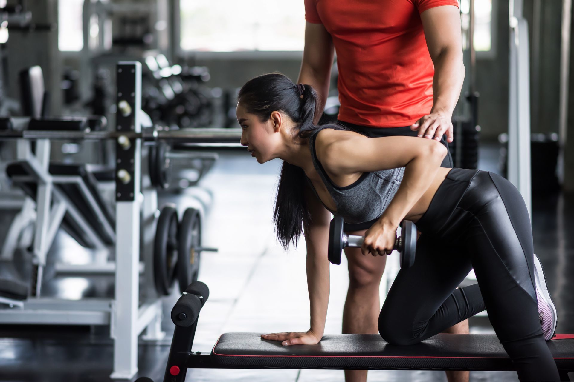 A man is helping a woman lift a dumbbell on a bench in a gym.