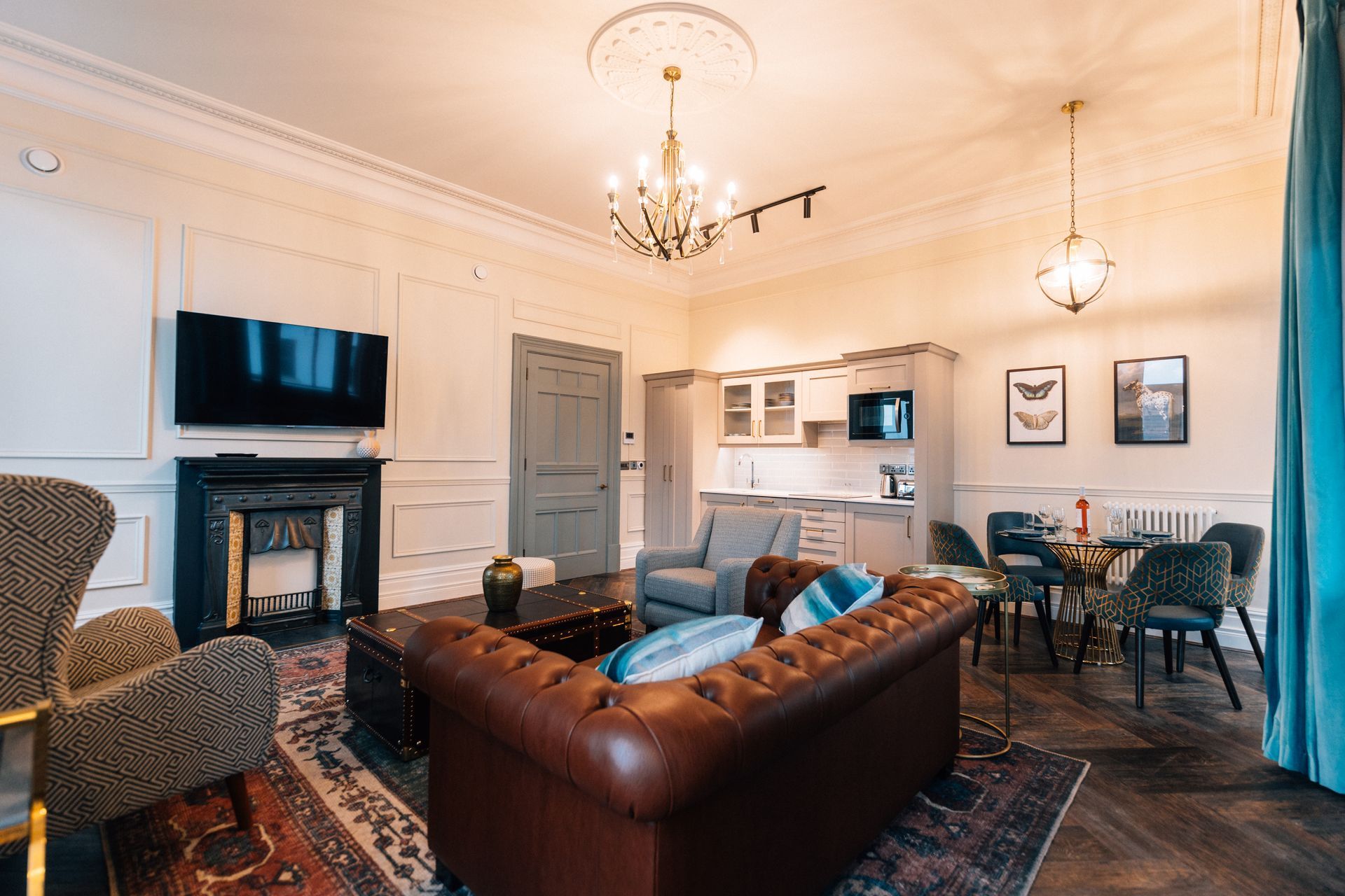 Elegant living room with brown leather sofa, fireplace, and open kitchen. Dark wood floors and ornate ceiling details.