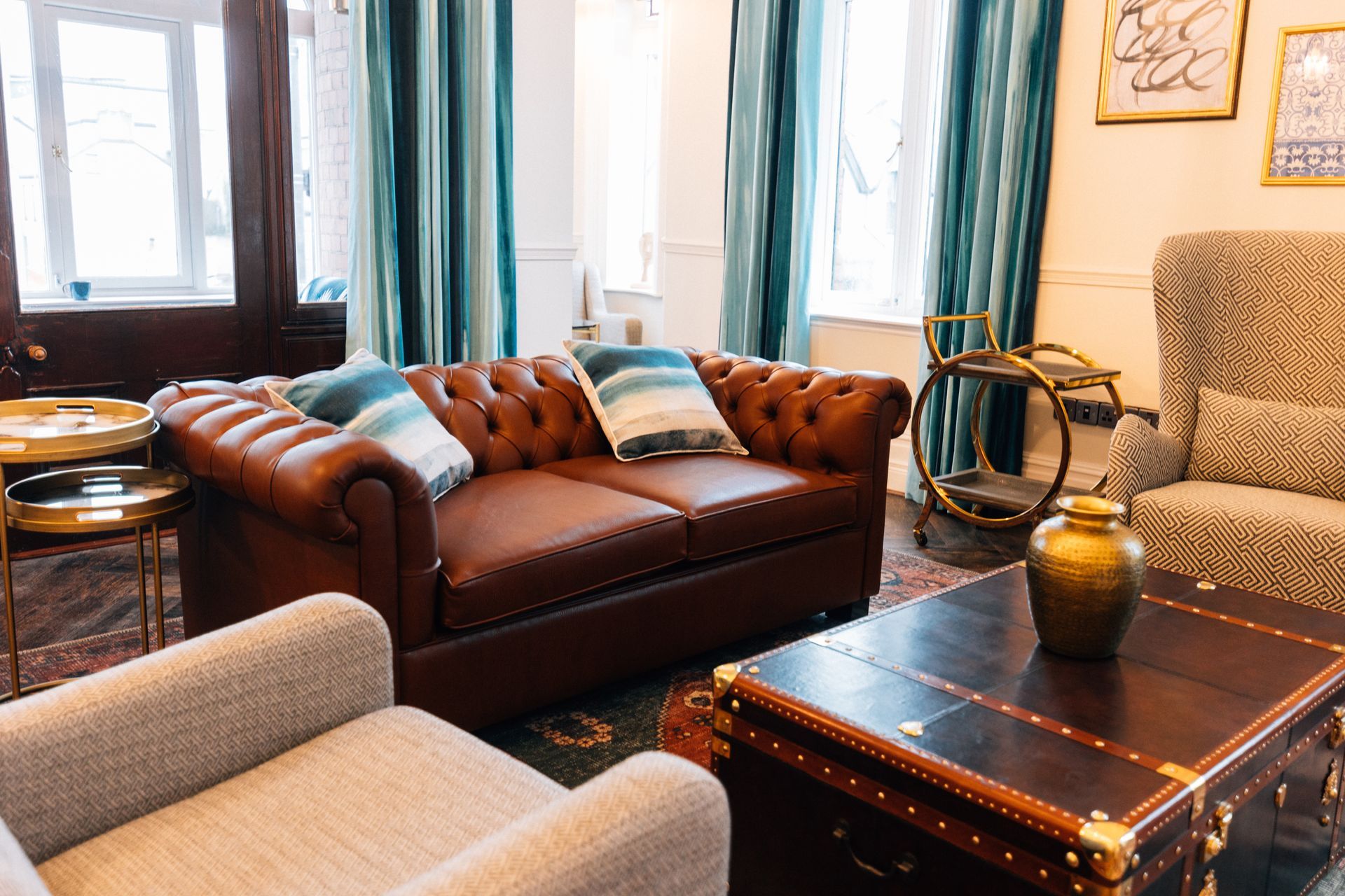 Cozy living room with brown leather sofa, patterned armchair, coffee table, teal curtains, and gold accents.