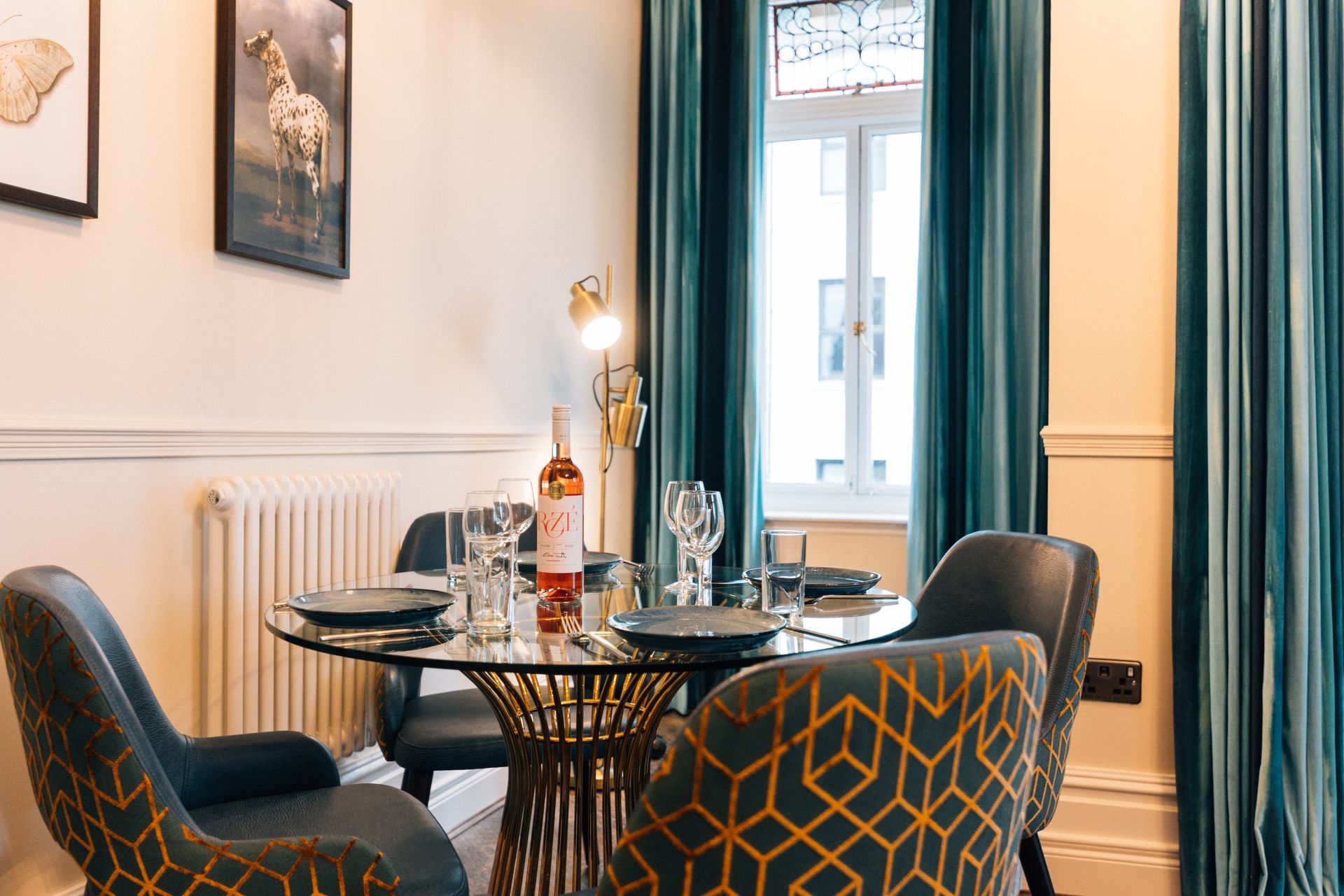 A dining room with a glass table, four chairs, teal curtains, and a bottle of rosé.