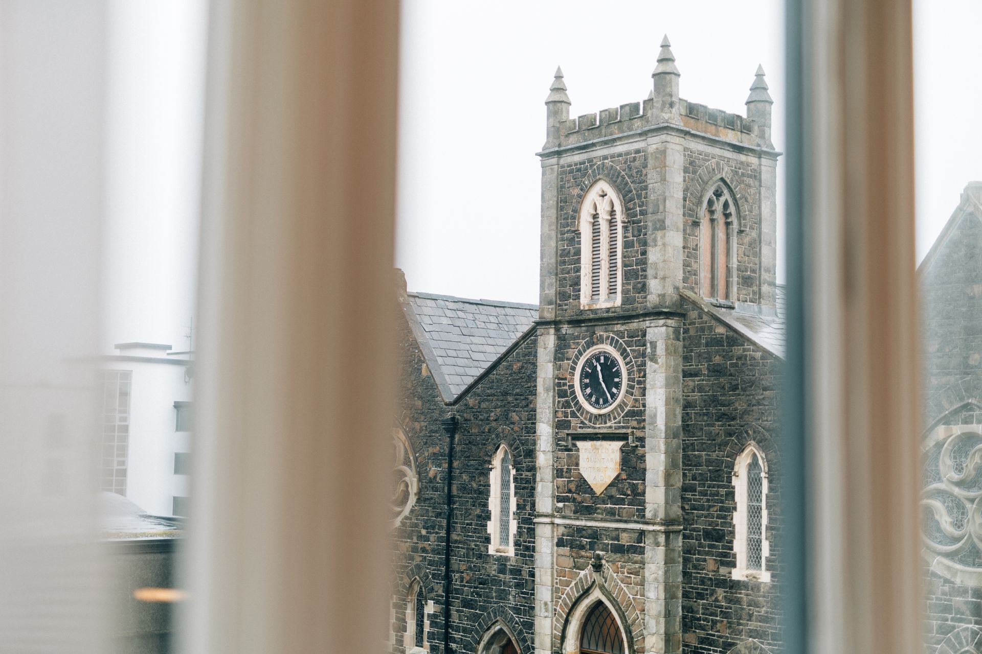 A stone church tower with a clock is seen through a window with white curtains, on an overcast day.