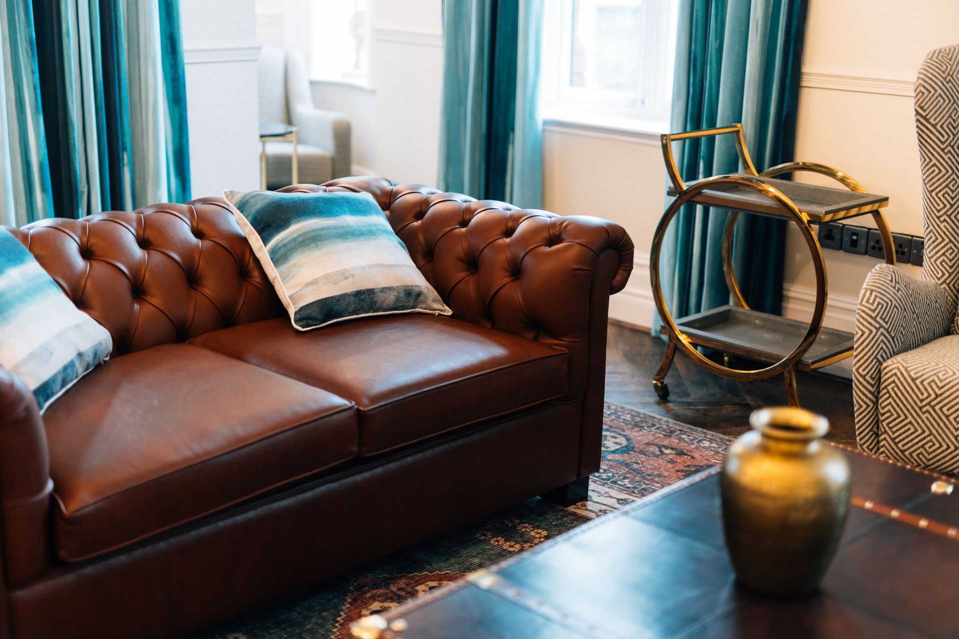 Leather sofa with blue pillows in a living room. Gold accents, teal curtains, and patterned rug.