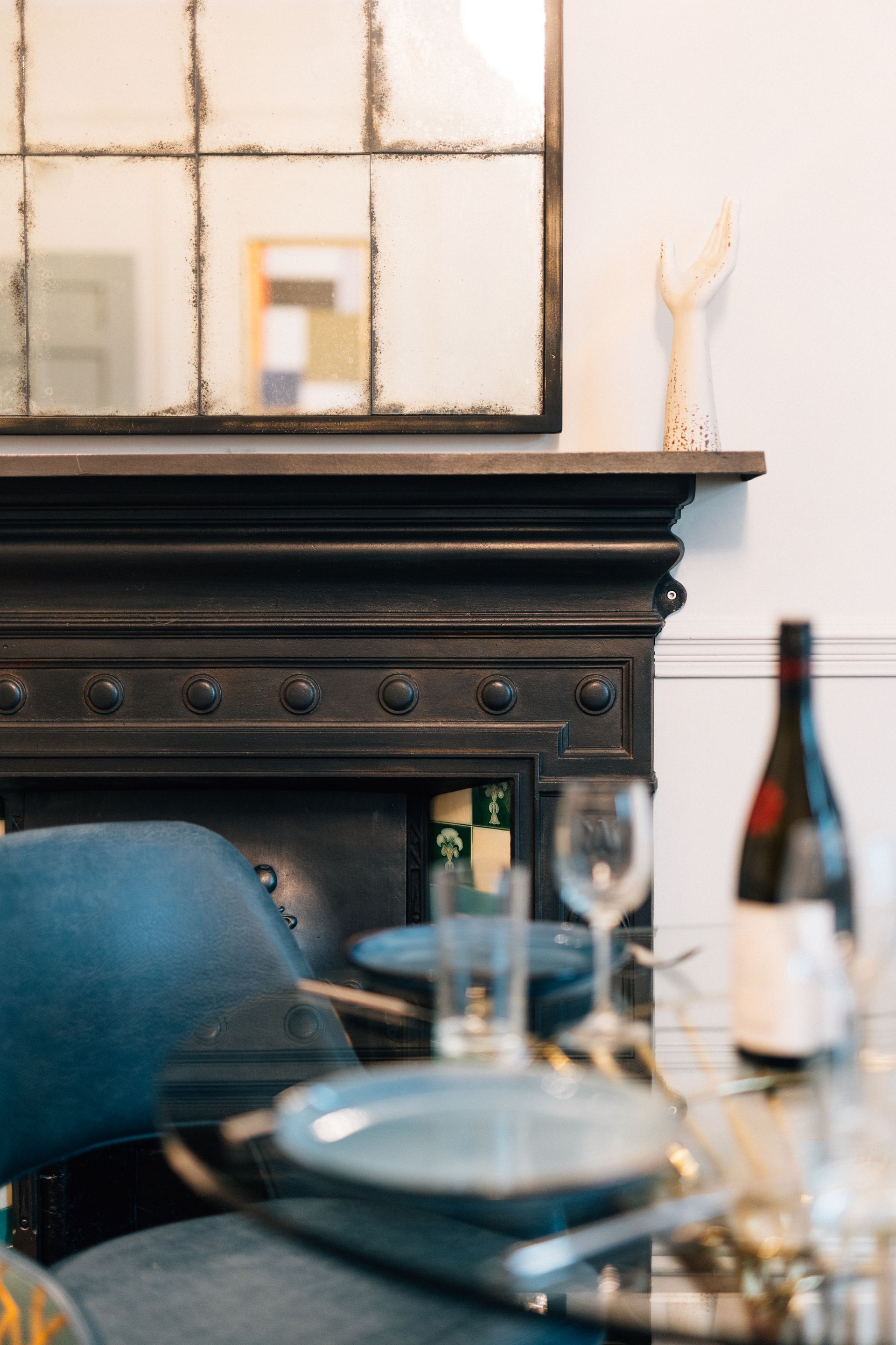 A black fireplace mantel with a mirror above and a table setting in front. A blue armchair is in the foreground.