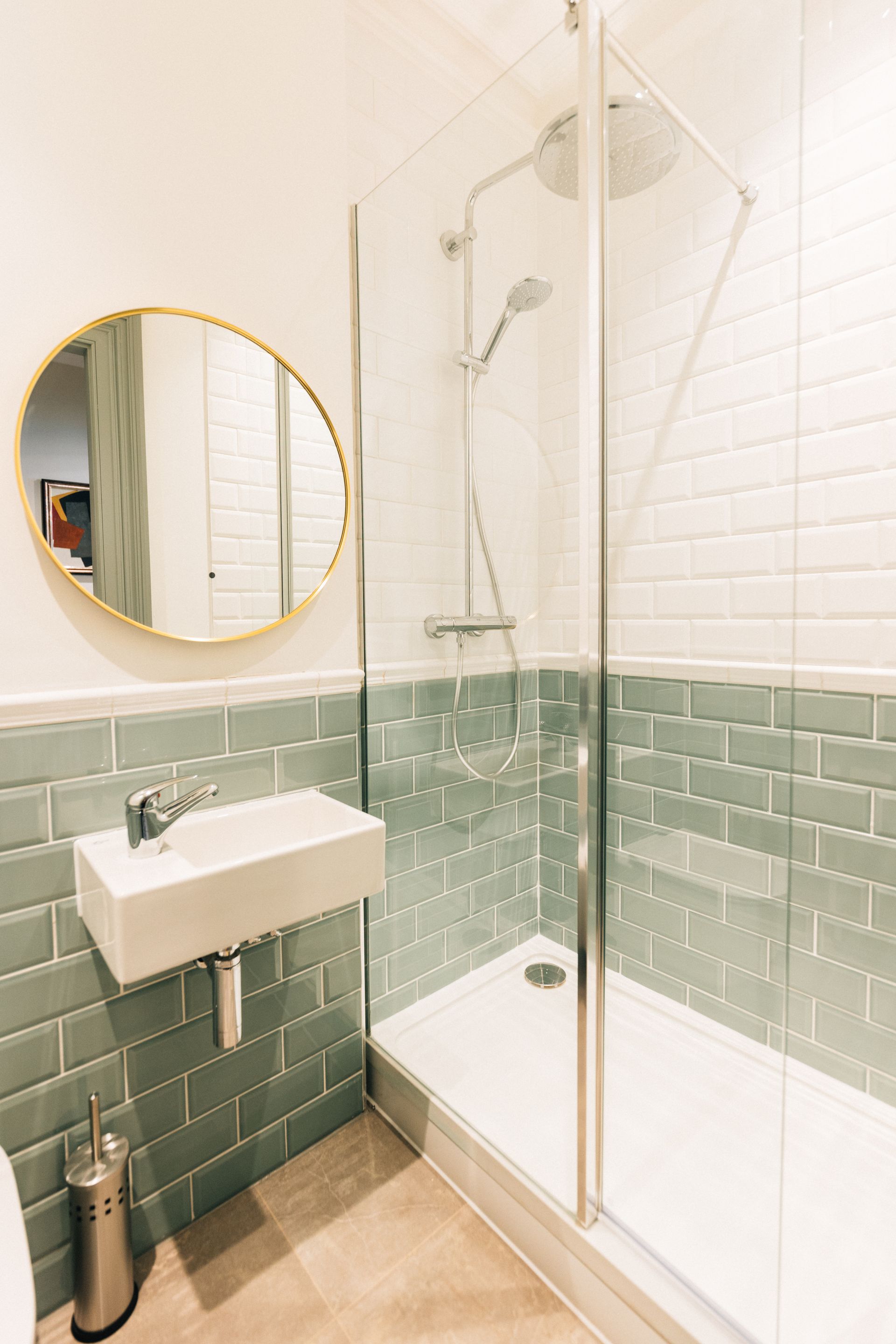 Small bathroom with a glass shower enclosure, a white sink, and a teal tiled wall. A round gold mirror is above the sink.