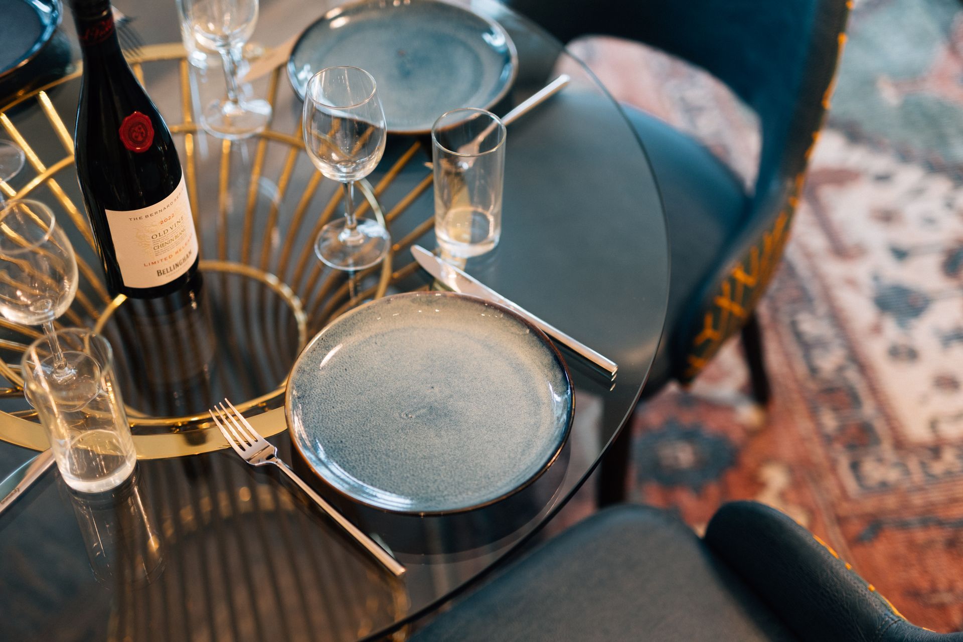 Table set for a meal with plates, glasses, silverware, and a wine bottle on a glass-topped table.