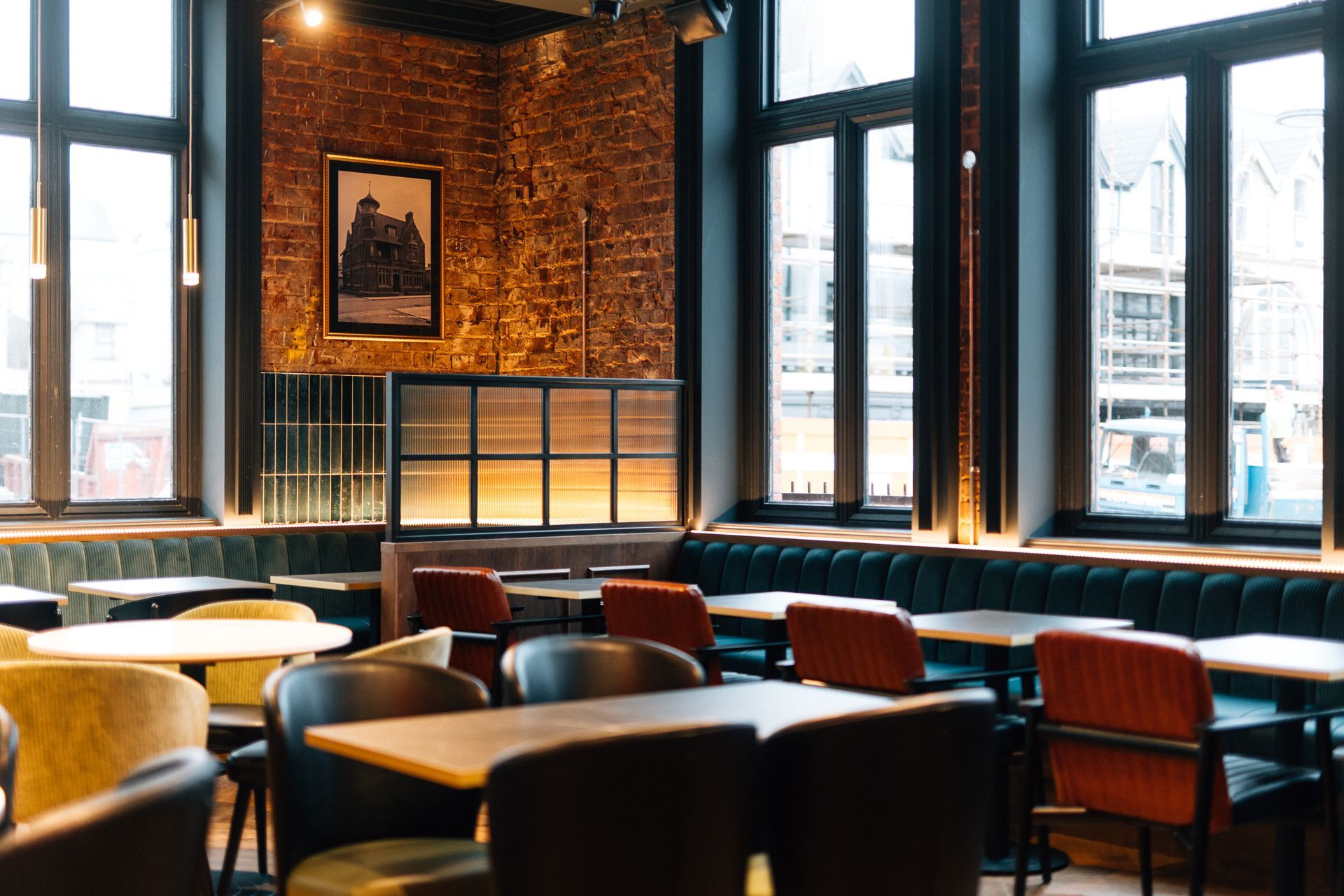 Interior of a restaurant with tables and chairs, a brick wall with a picture, and large windows overlooking a street.