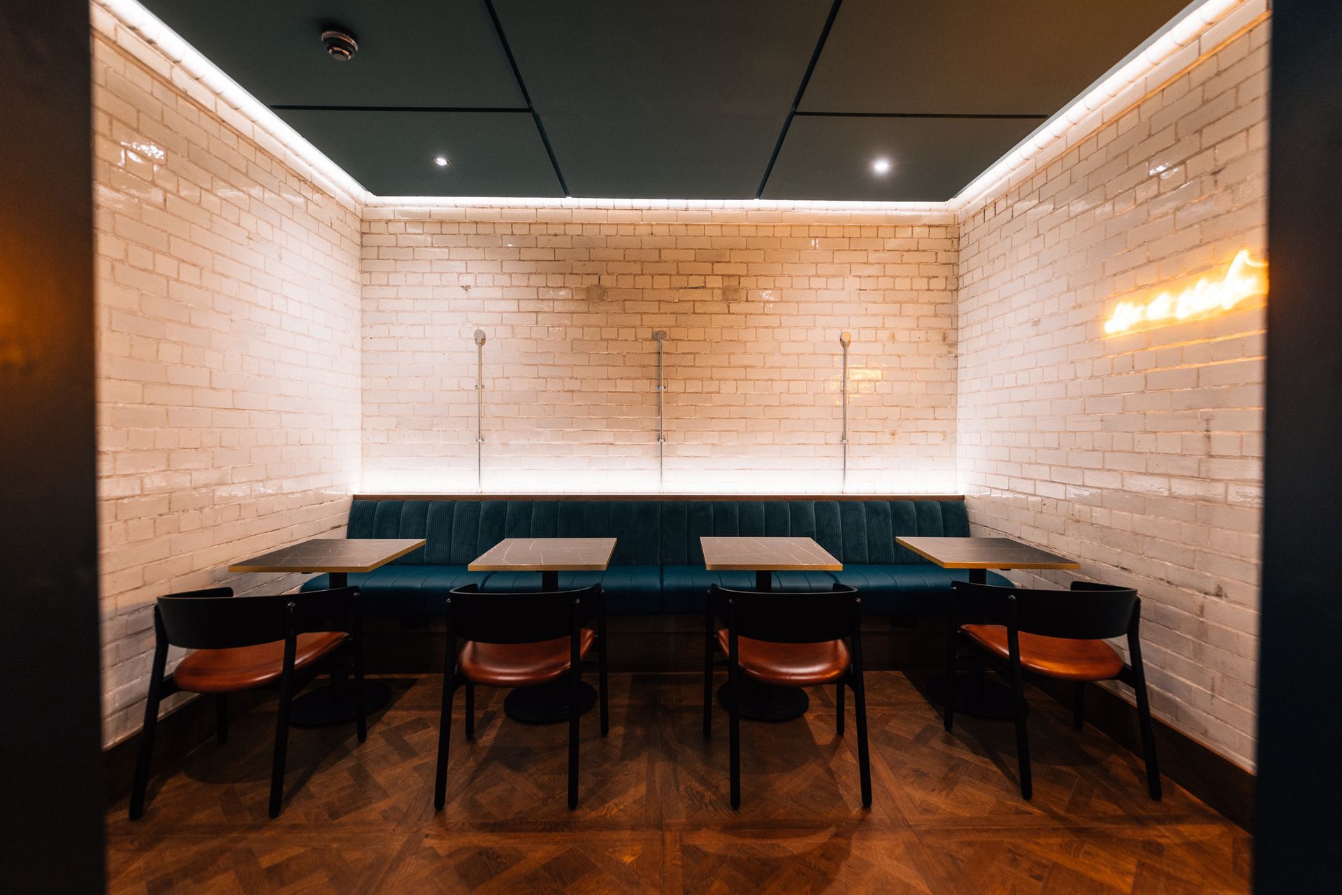 Cozy booth seating in a restaurant.  White brick walls surround a teal bench, tables, and dark chairs.  A neon sign glows.