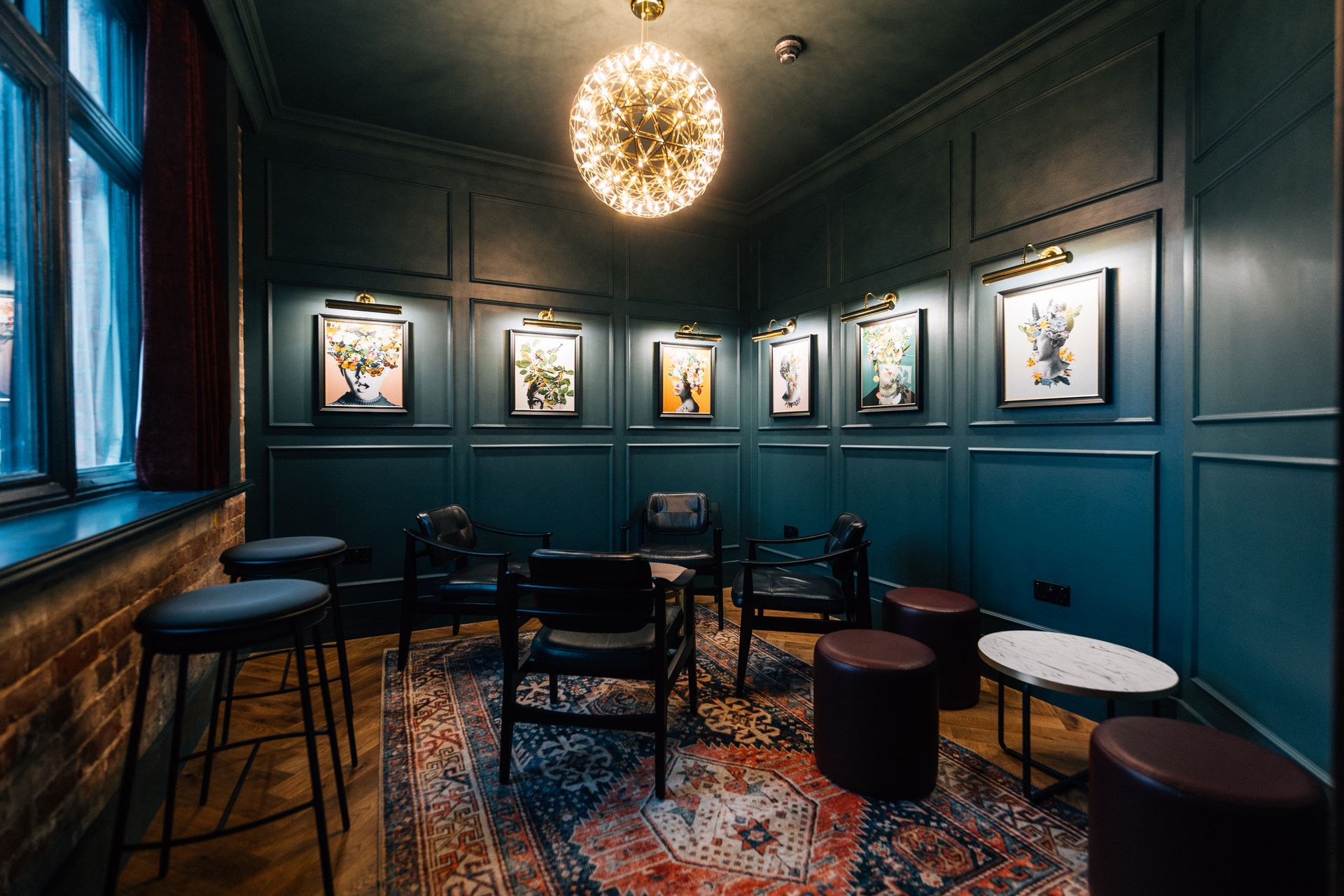 A dimly lit lounge with teal panelled walls, art, and seating including stools, chairs, and a rug.