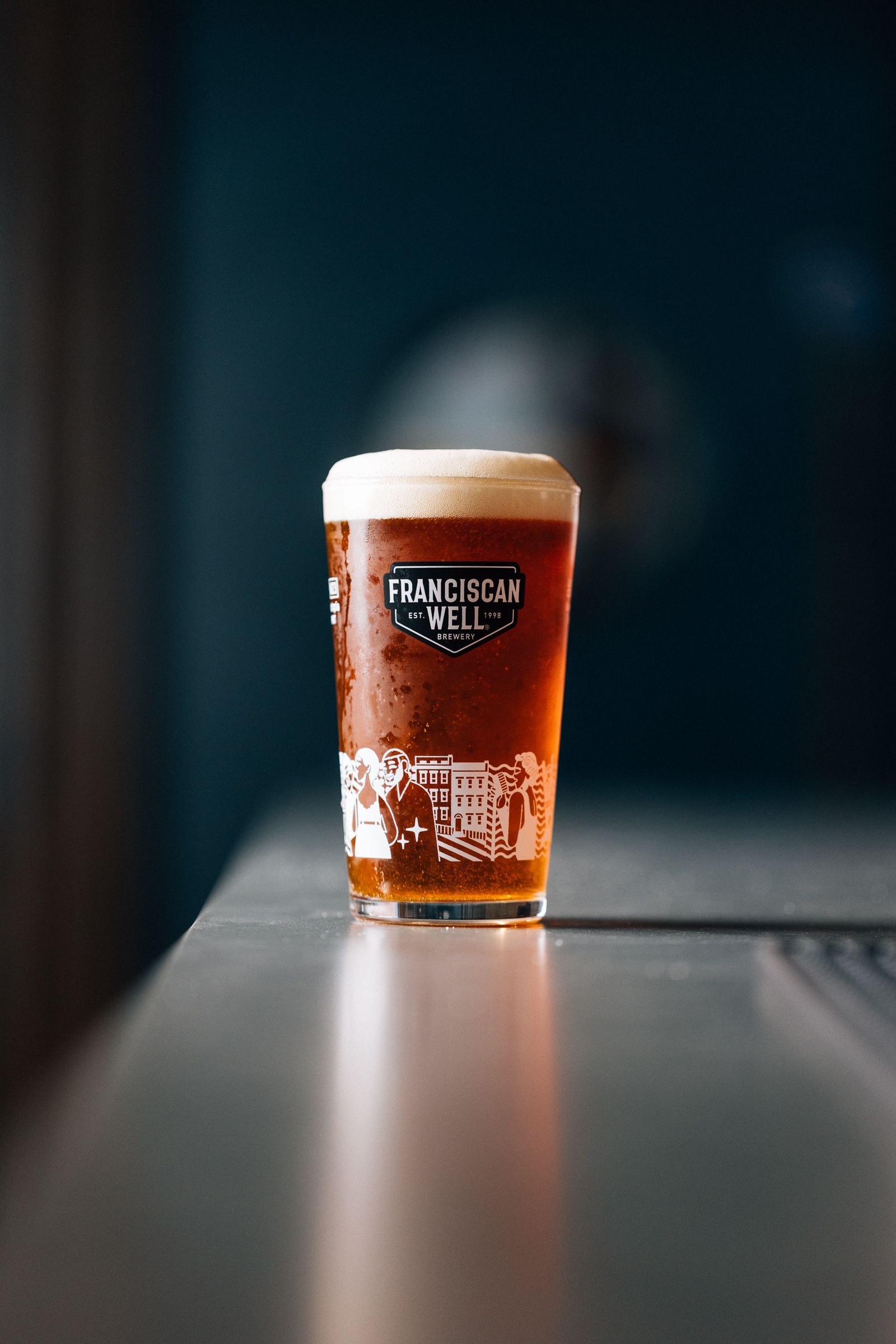 A pint of dark beer with a foamy head sits on a bar. The glass has a logo and is slightly frosted.