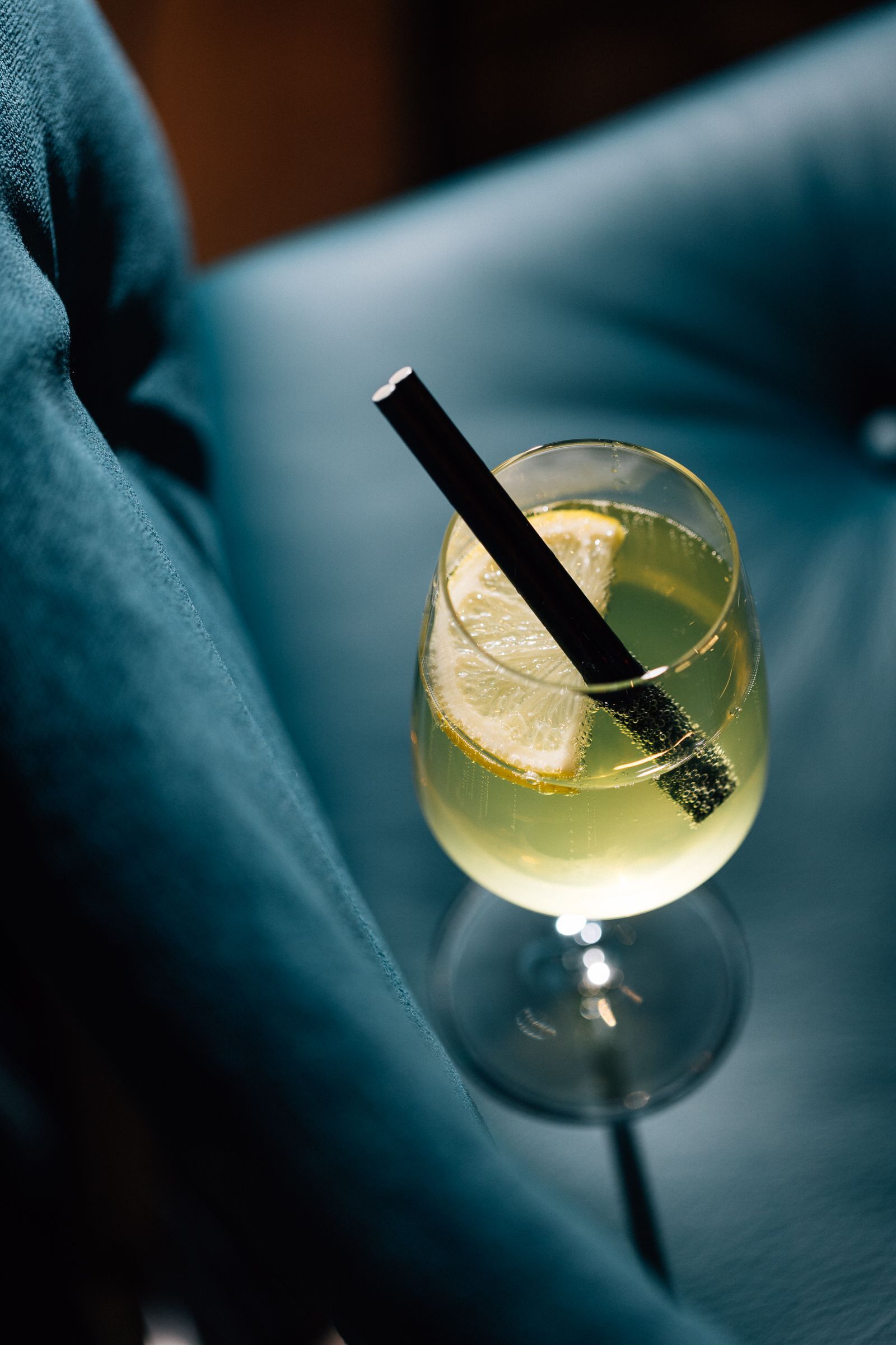 A glass of pale yellow cocktail with a lemon slice and a black straw, resting on a dark surface.