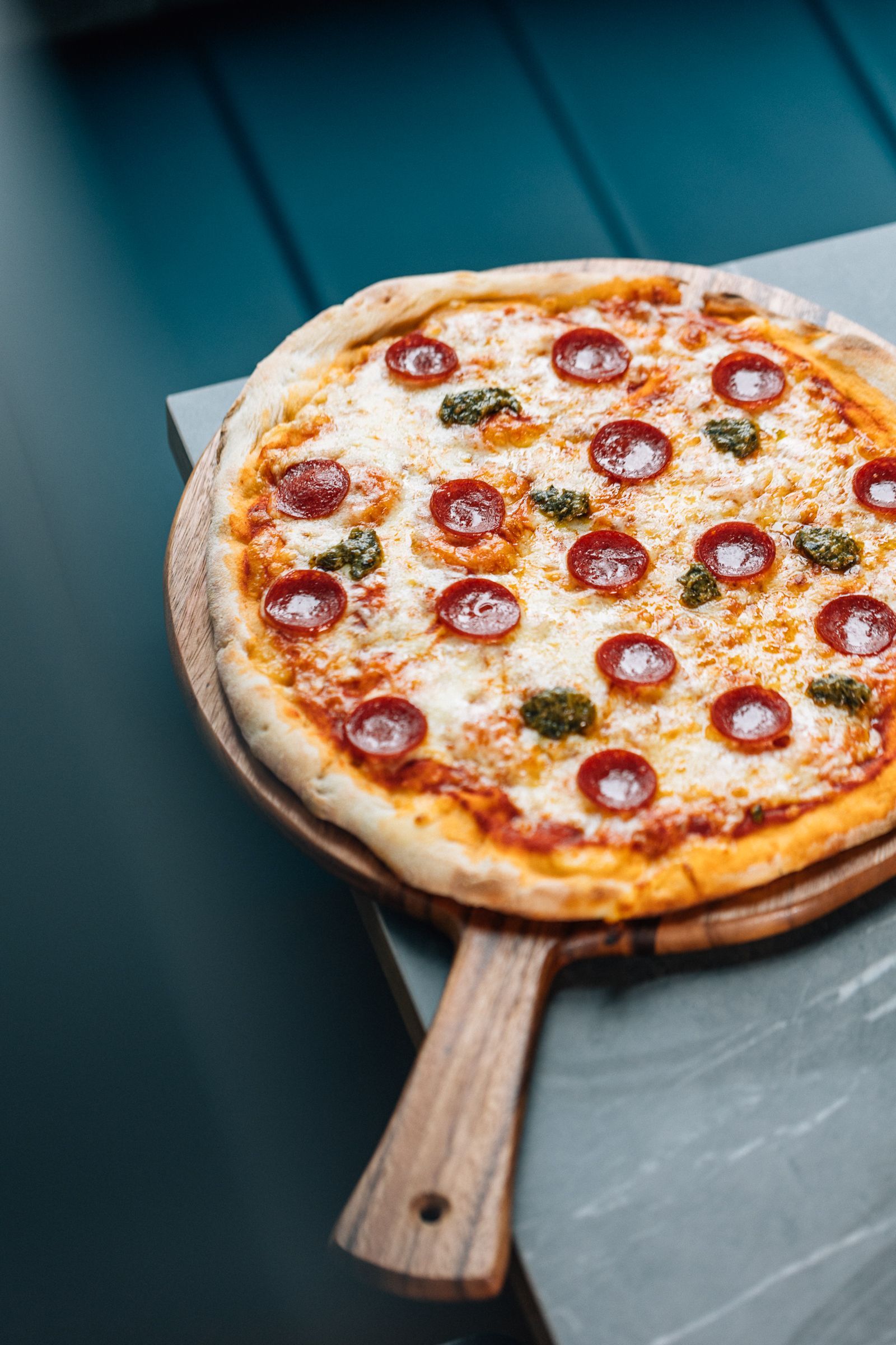 Pepperoni pizza on a wooden serving board against a teal backdrop. 
