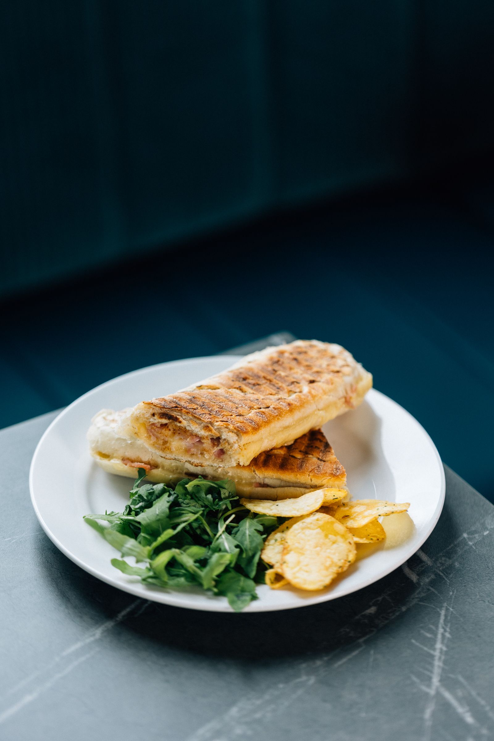 Grilled sandwich on a white plate with arugula and potato chips, set against a dark blue backdrop.