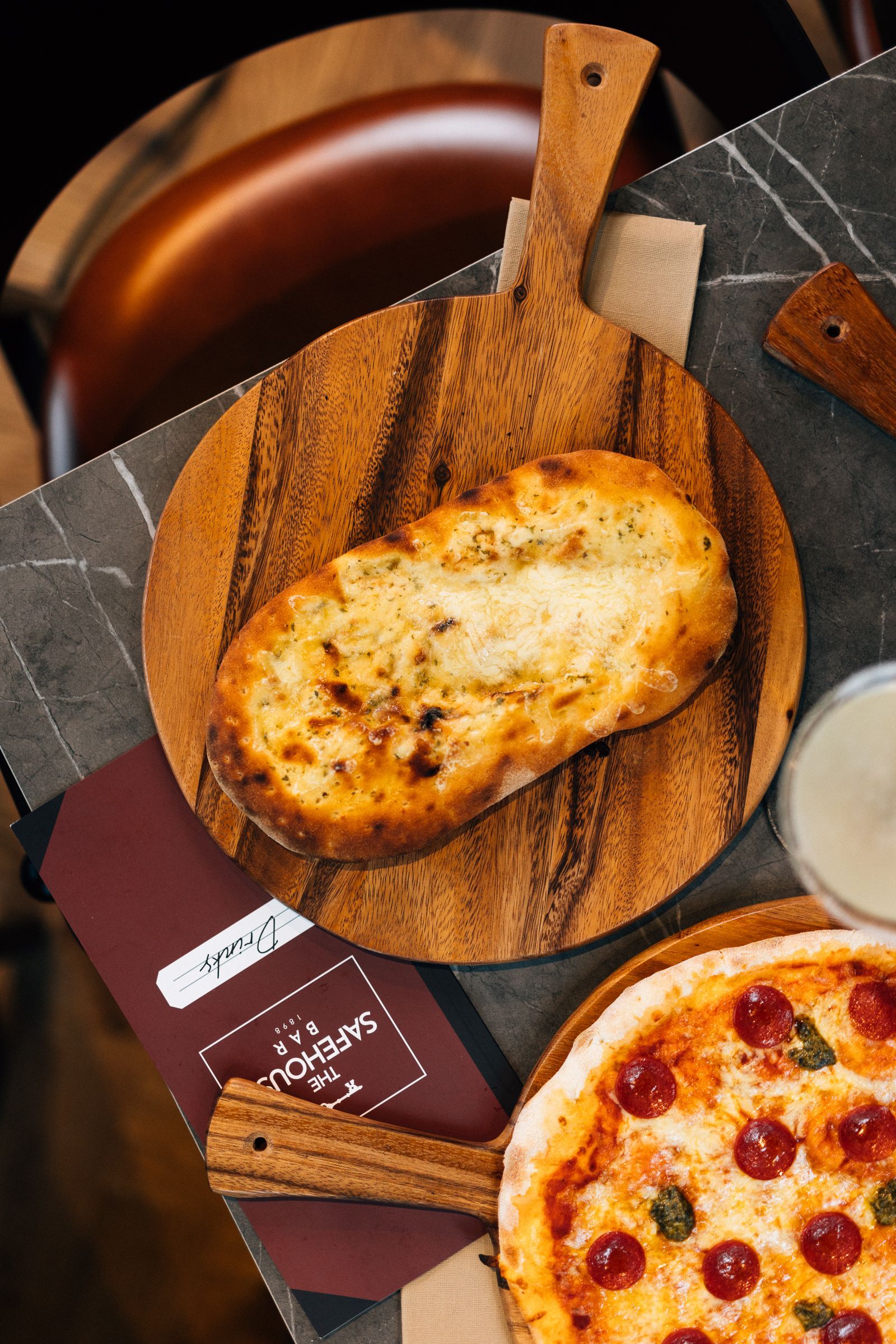 A toasted garlic bread slice on a wooden cutting board, with a pizza below and a dipping sauce.