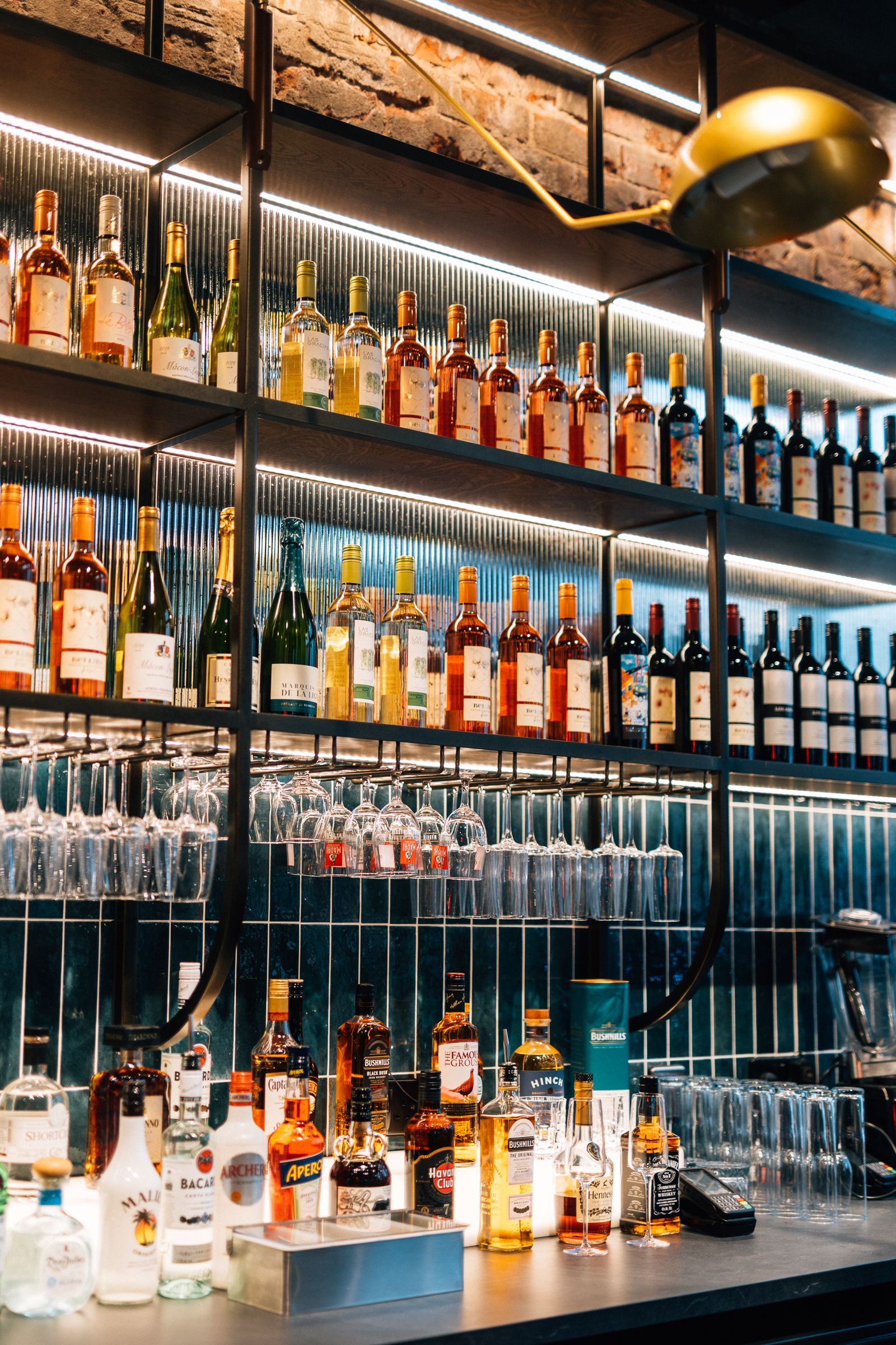 Bar interior with shelves stocked with bottles and hanging glassware, lit by rows of lights.