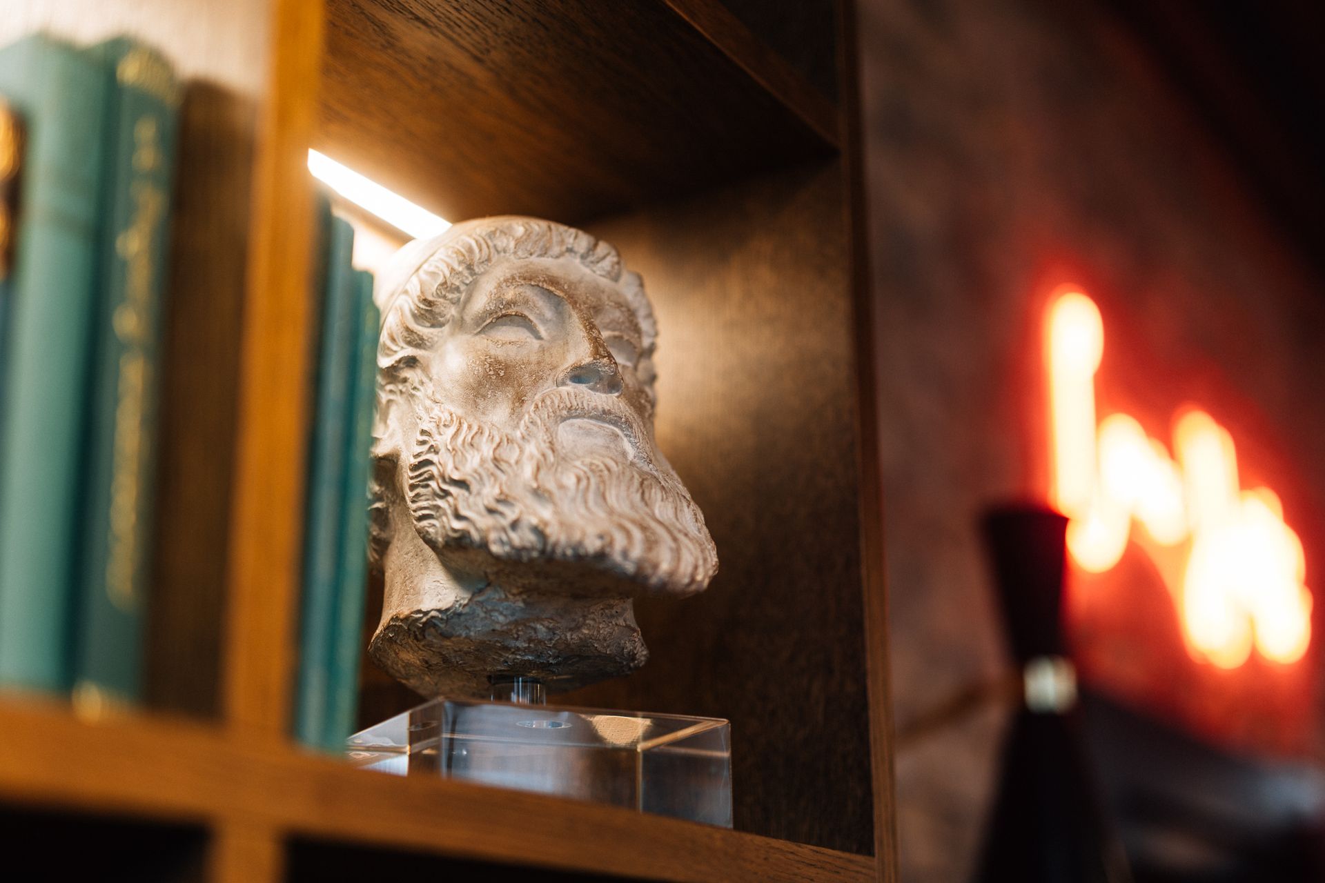 A marble bust of a bearded man on a shelf, books to the left, and a blurred neon sign to the right.