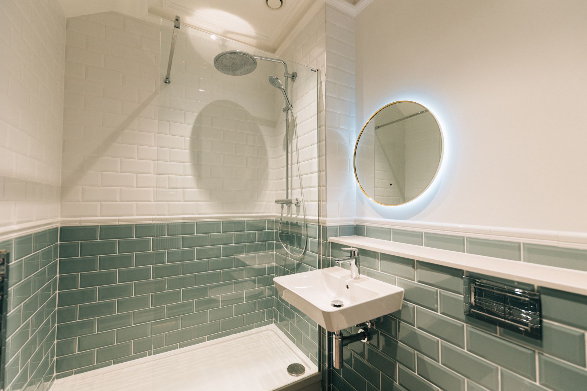 Small bathroom with gray and white tile walls, glass shower, round backlit mirror, and wall-mounted sink.