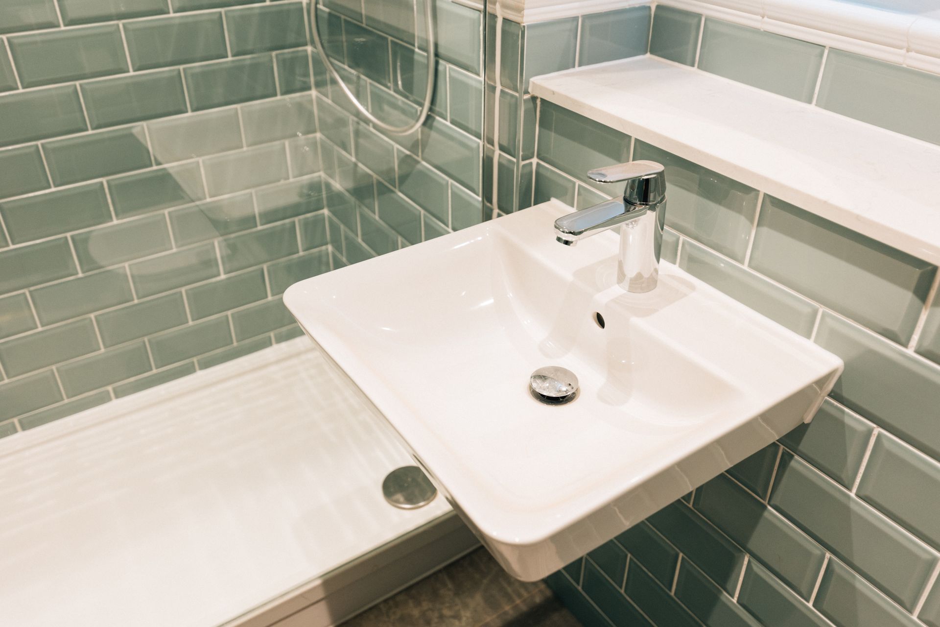 Small white bathroom sink with chrome faucet against green tiled wall. A shower is to the left.