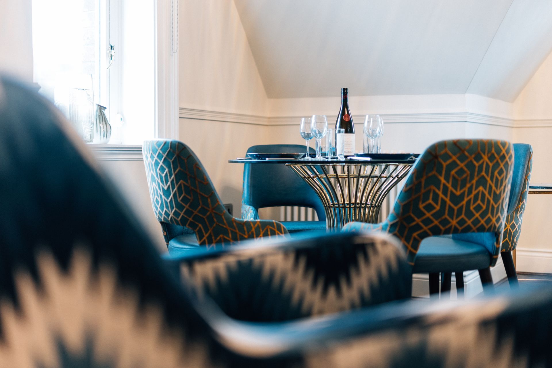 Dining area with blue patterned chairs around a round table set for a meal. A wine bottle and glasses are on the table.