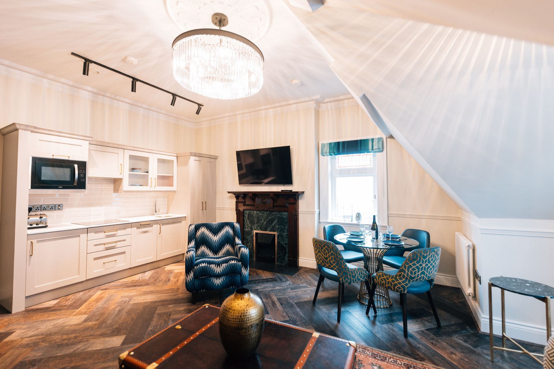 Cozy hotel suite with a kitchen, seating area, and small dining table; beige walls, wooden floors, and a crystal chandelier.