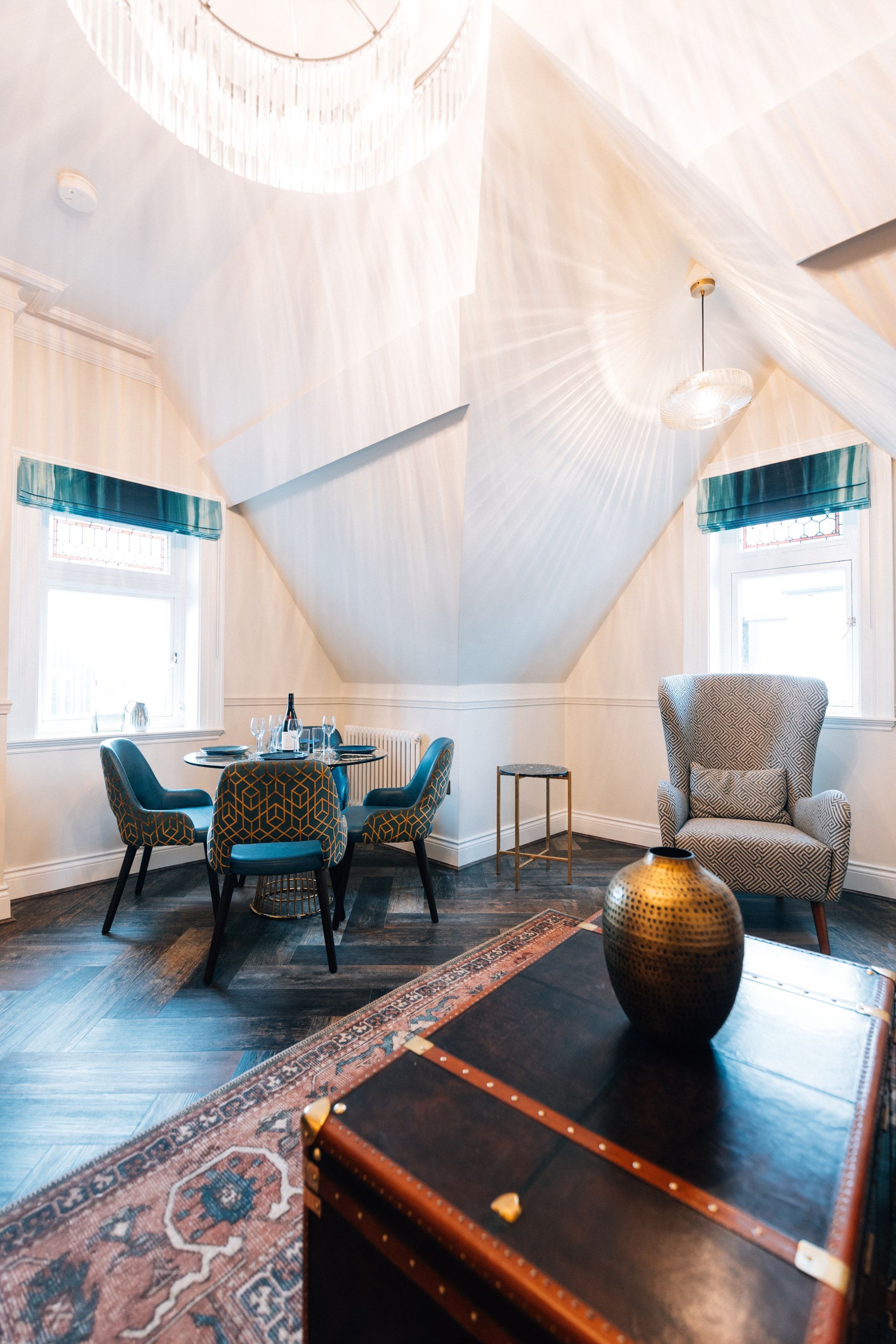 Features a table with blue patterned chairs, a patterned armchair, and a trunk with a golden vase on a patterned rug.