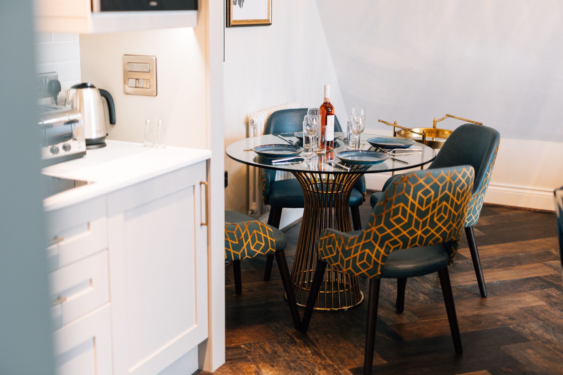Dining area with a round glass table, four chairs with patterned upholstery, and a small kitchen visible in the foreground.