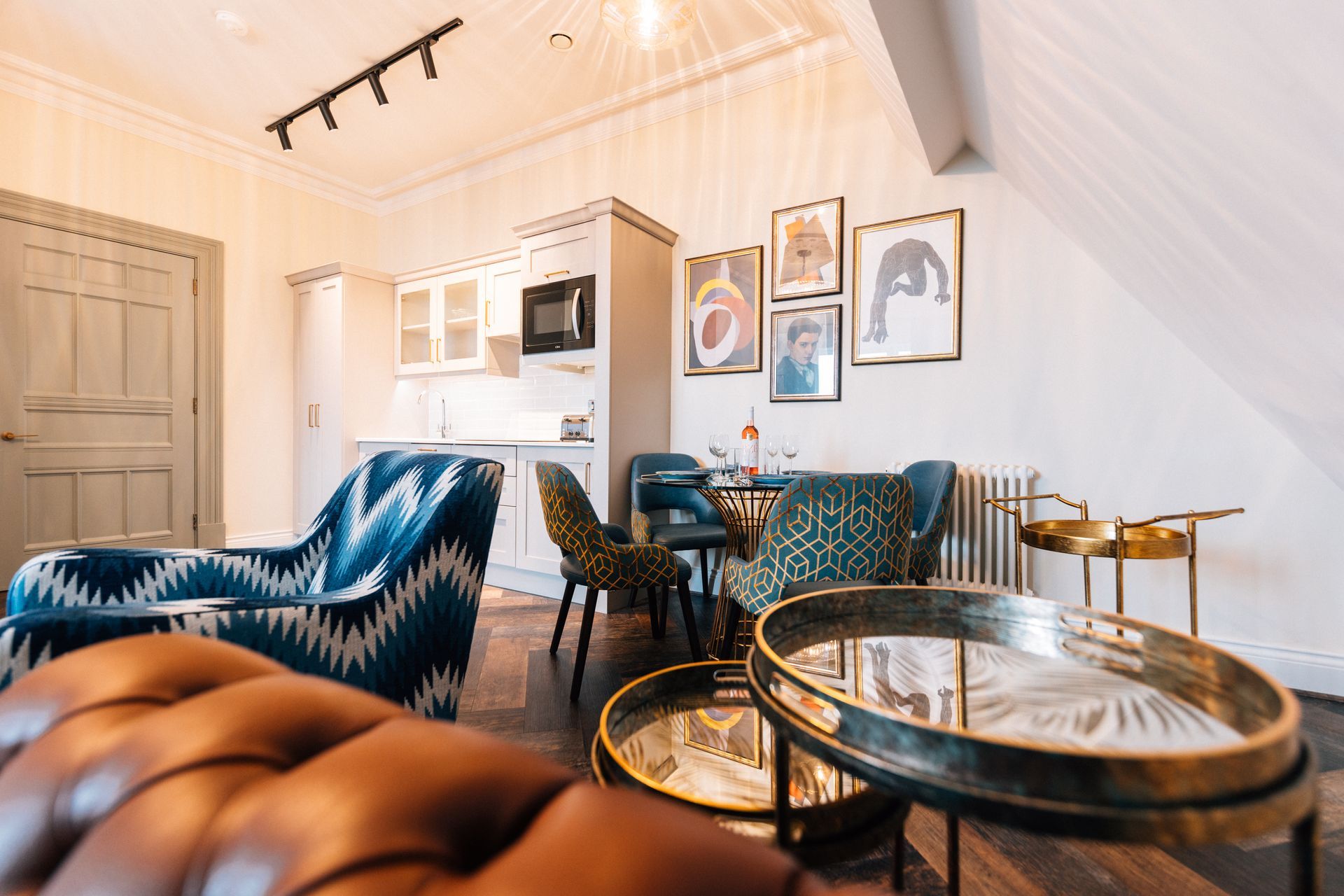 Living room with a kitchenette. Features a blue patterned sofa, a leather couch, round tables, and art on the wall.