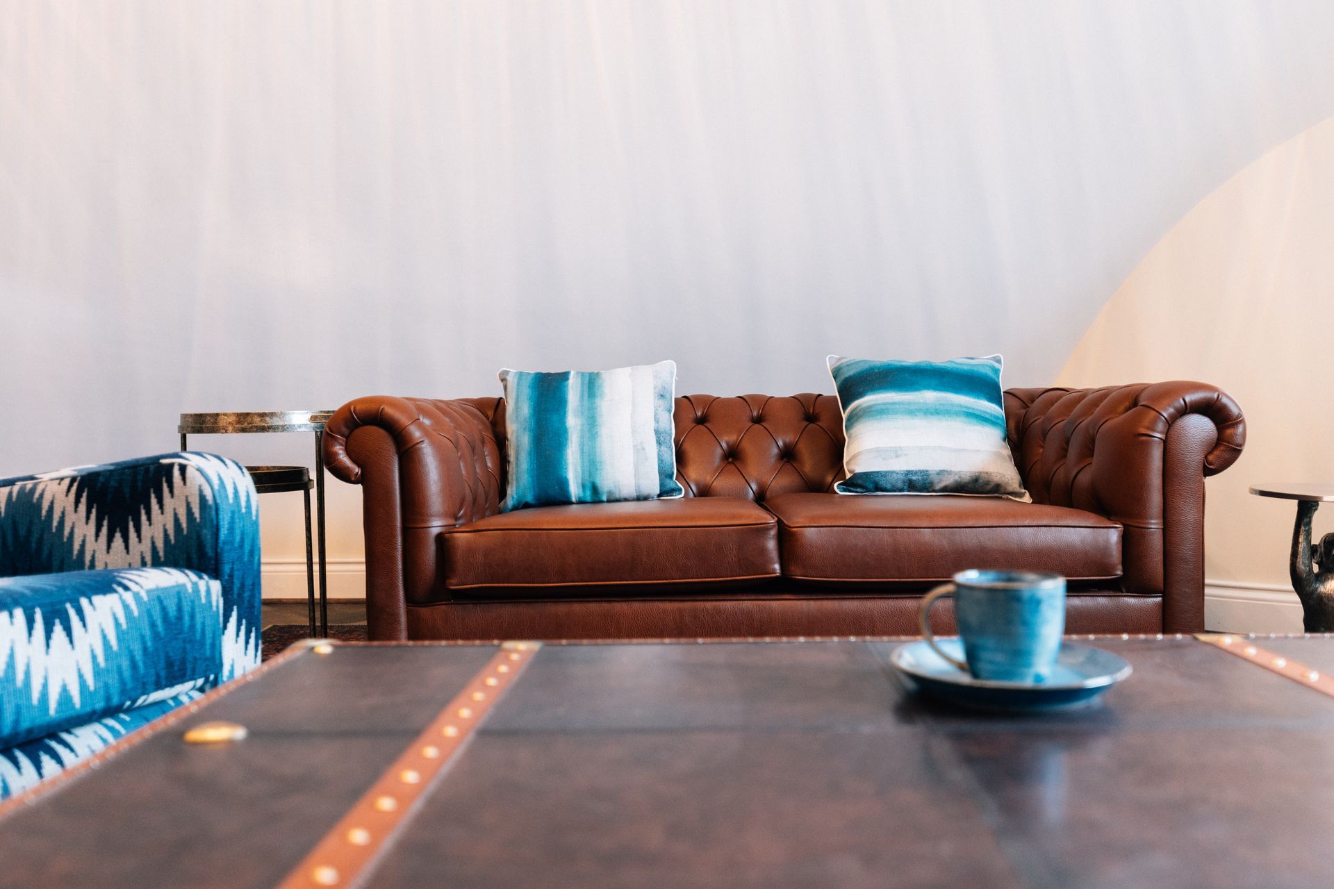 Living room with brown leather sofa, blue pillows, and a patterned armchair; coffee table with a teacup.